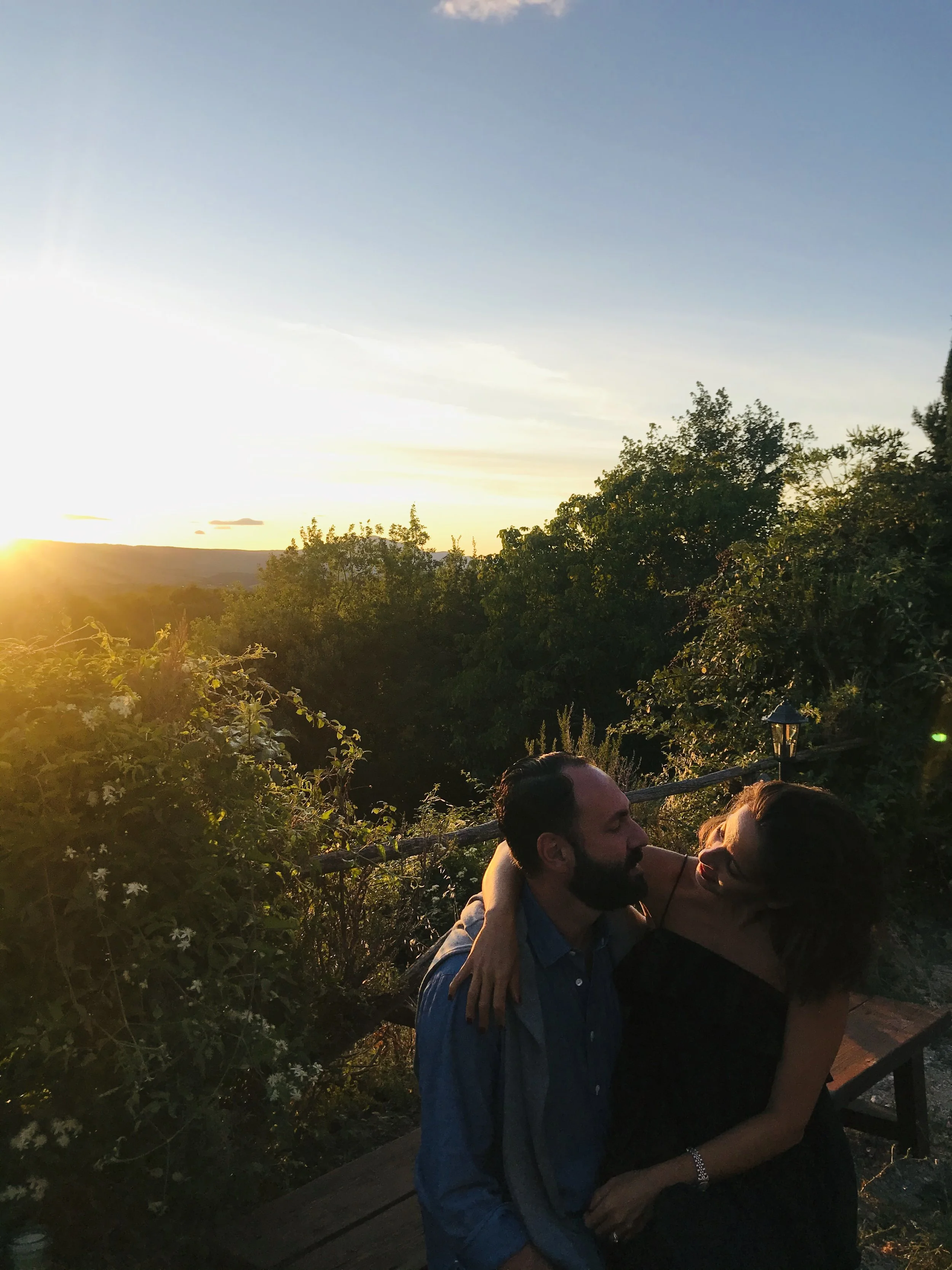 Due persone, un uomo e una donna, abbracciati e sorridenti, si stanno guardando con affetto in un ambiente esterno al tramonto, circondati da vegetazione e alberi, con il cielo che si tinge di colori caldi.