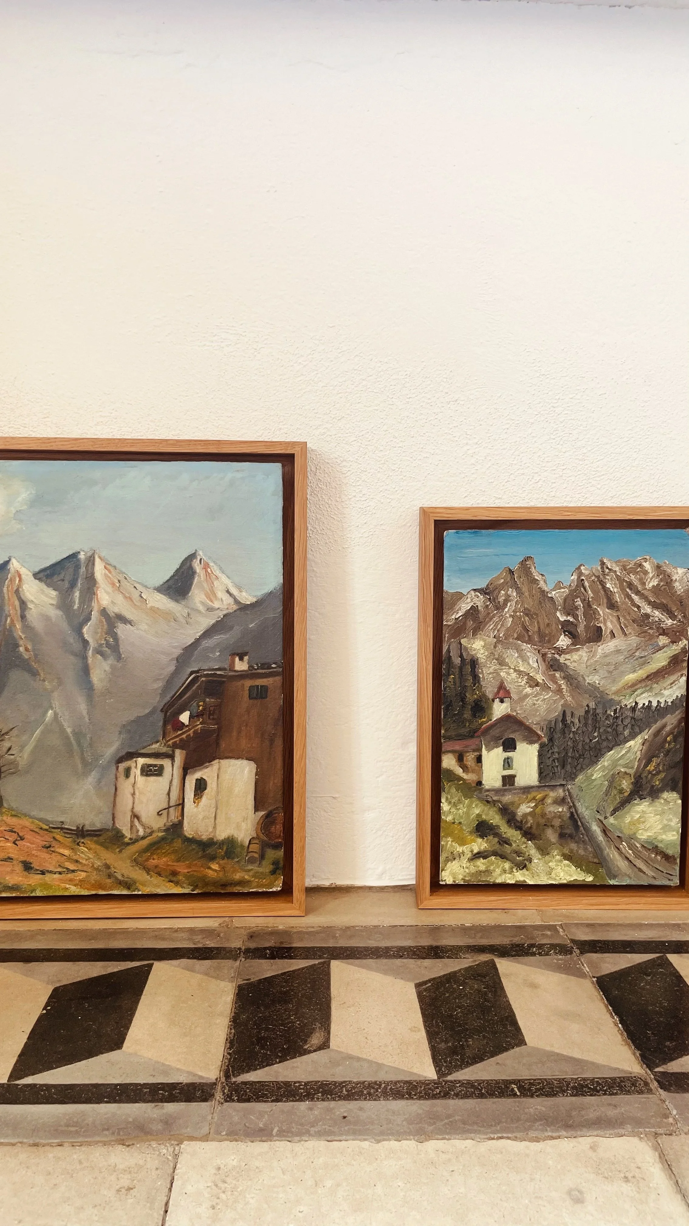 Due dipinti che raffigurano paesaggi montani con case, con una cornice di legno, appesi a una parete bianca. Sul pavimento c'è un disegno in piastrelle con un motivo geometrico.