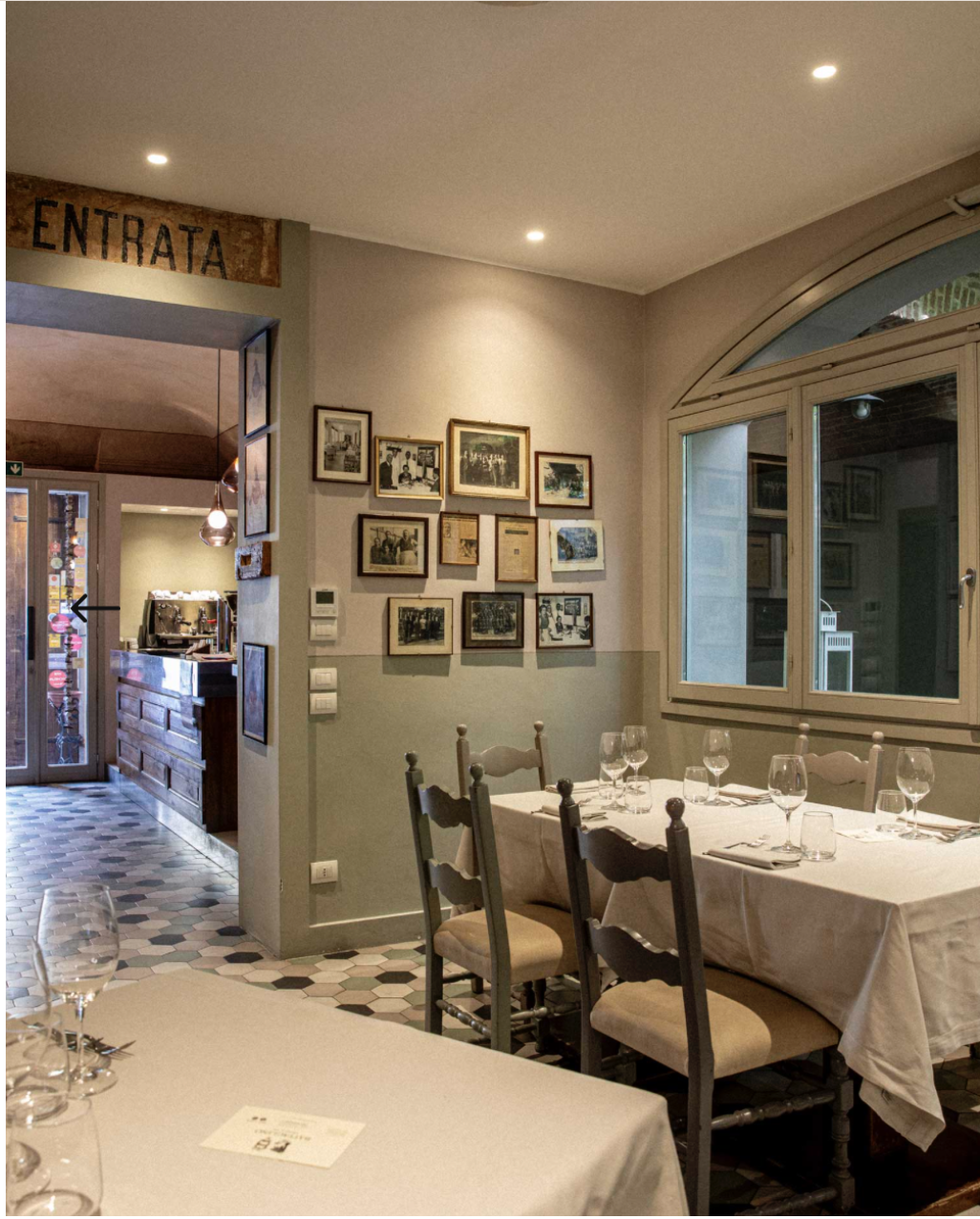 Interno di un ristorante con parete decorata con fotografie in cornice, tavolo apparecchiato con bicchieri e posate, grande finestra e segnale 'Entrata' sopra l'ingresso.