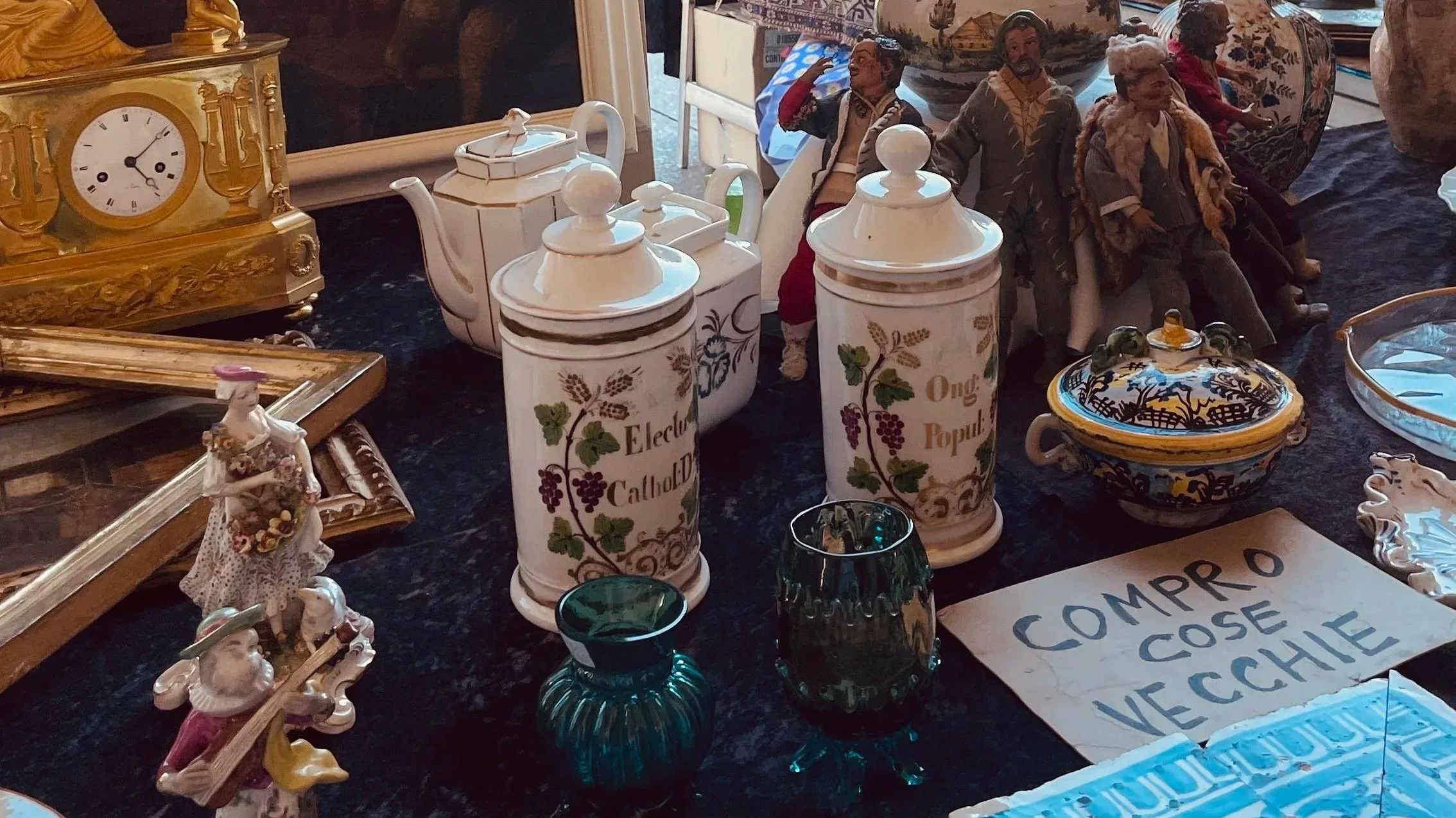 Varie oggetti d'antiquariato e decorativi su un tavolo, tra cui orologio a pendolo dorato, statuette raffiguranti persone, piatti decorativi e barattoli dipinti con scritte come "Elettro Caldob" e "Ong Popu". C'è anche un cartello che dice 'COMPRO COSE VECCHIE'.