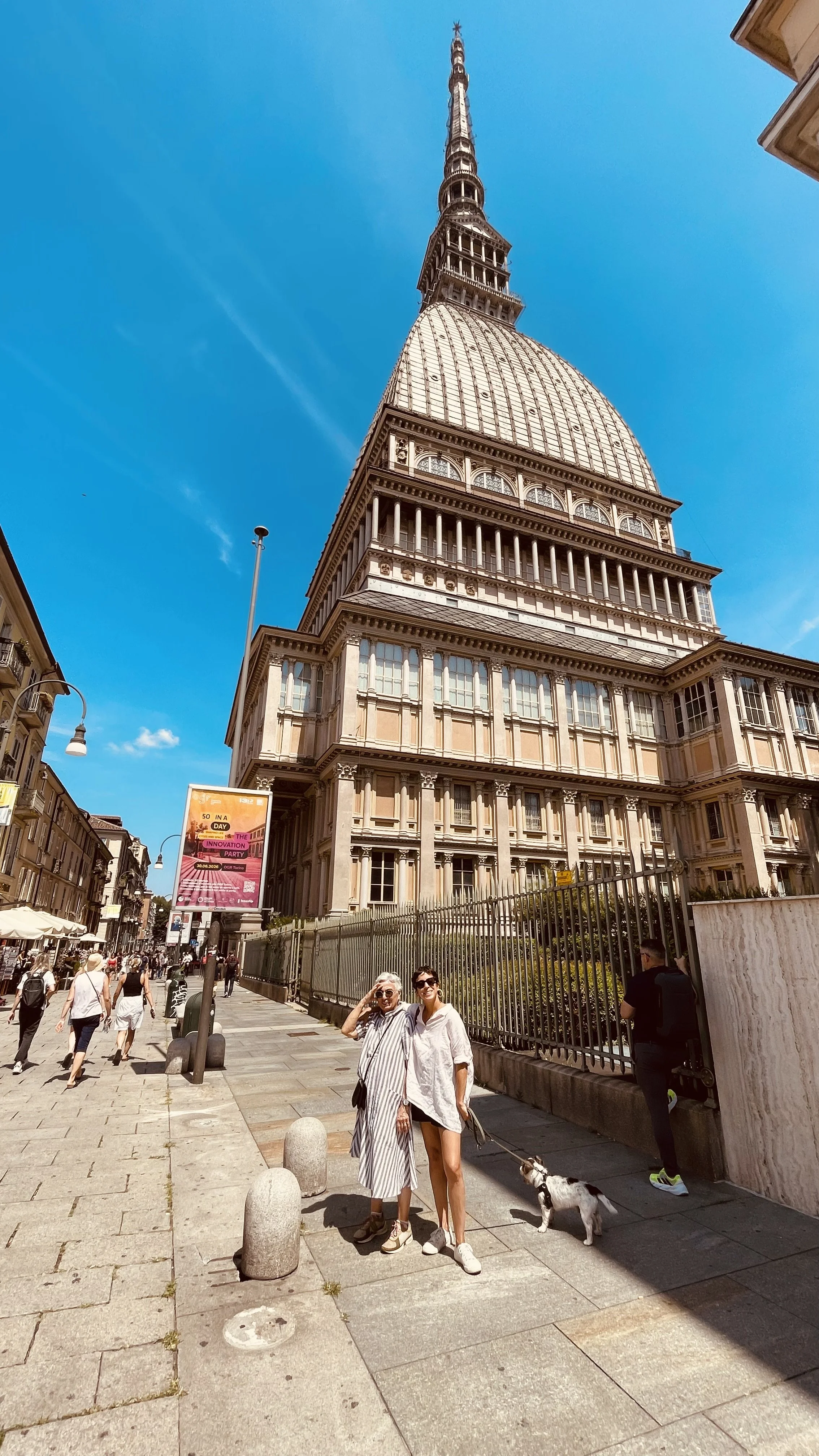 Due donne sorridenti in piedi su una strada affollata di fronte al famoso edificio Mole Antonelliana, a Torino, sotto un cielo sereno e blu.
