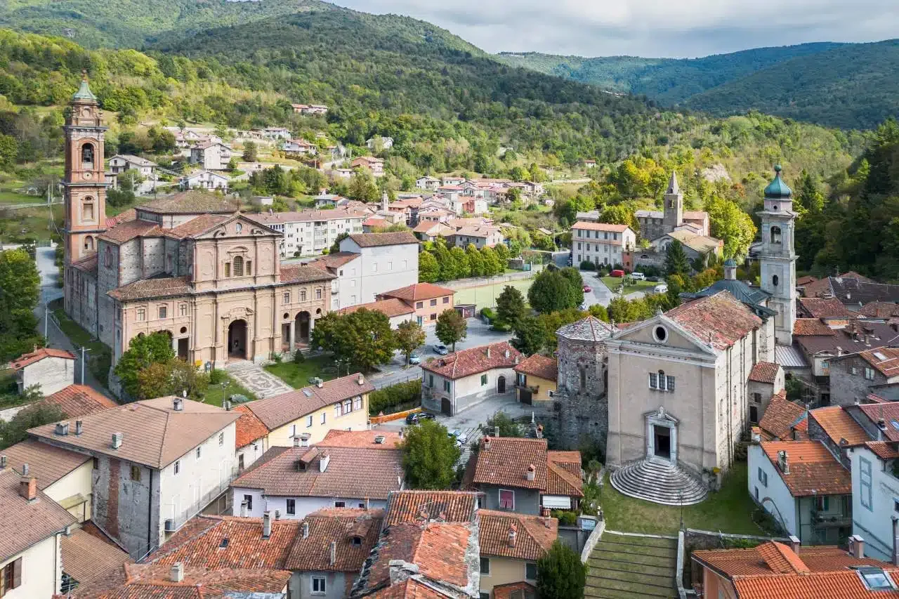 Vista di un piccolo paese in montagna con chiese storiche e case con tetti di tegole rosse, circondato da colline verdi e foreste.