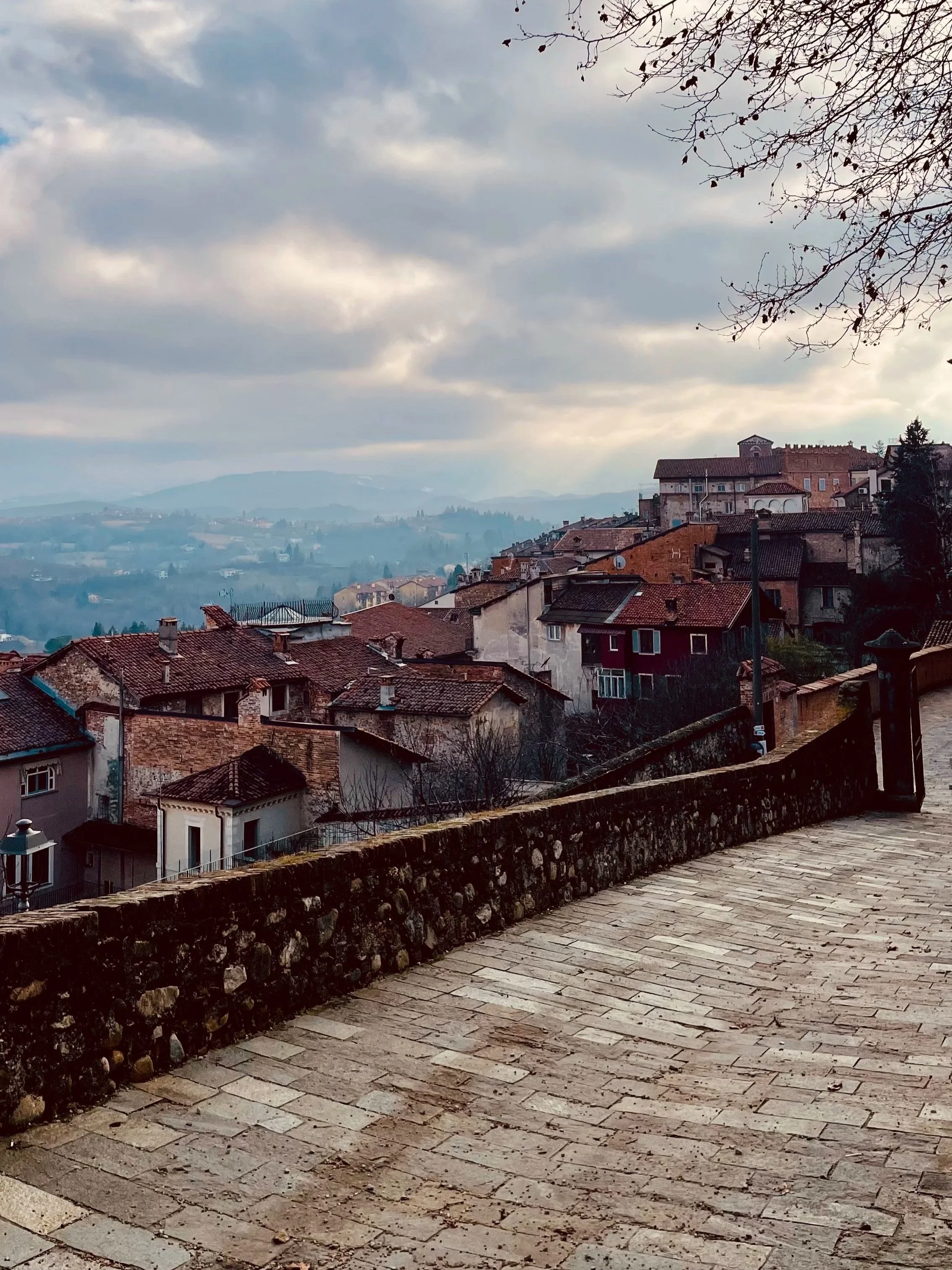 Veduta di un borgo antico concase in pietra e tetti di terracotta, sullo sfondo colline e montagne avvolte da una leggera foschia, cielo nuvoloso di giorno