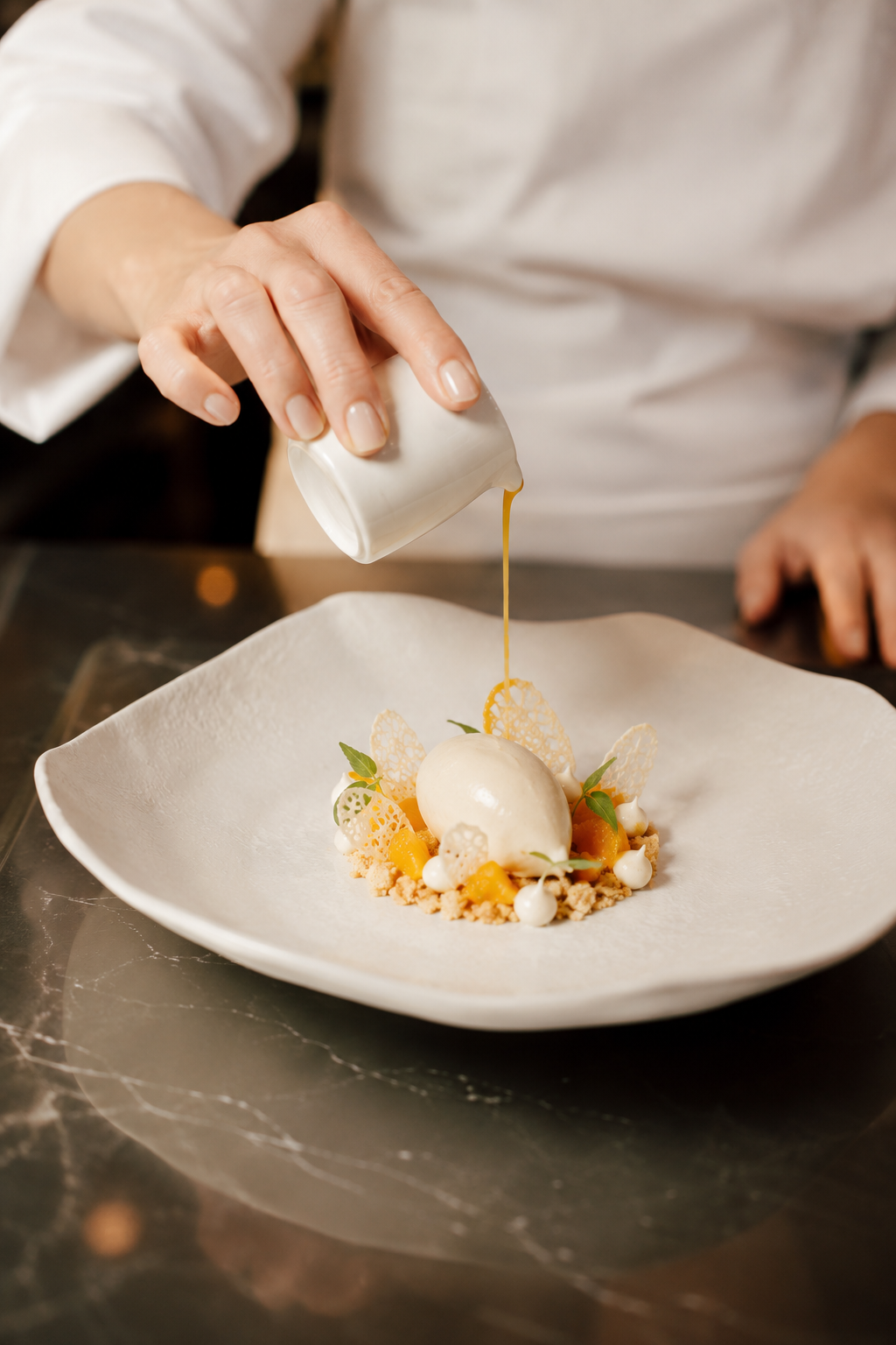 A chef decorates a dessert with sauce from a small pitcher, featuring a scoop of ice cream, fruit pieces, and decorative edible elements on a white plate.