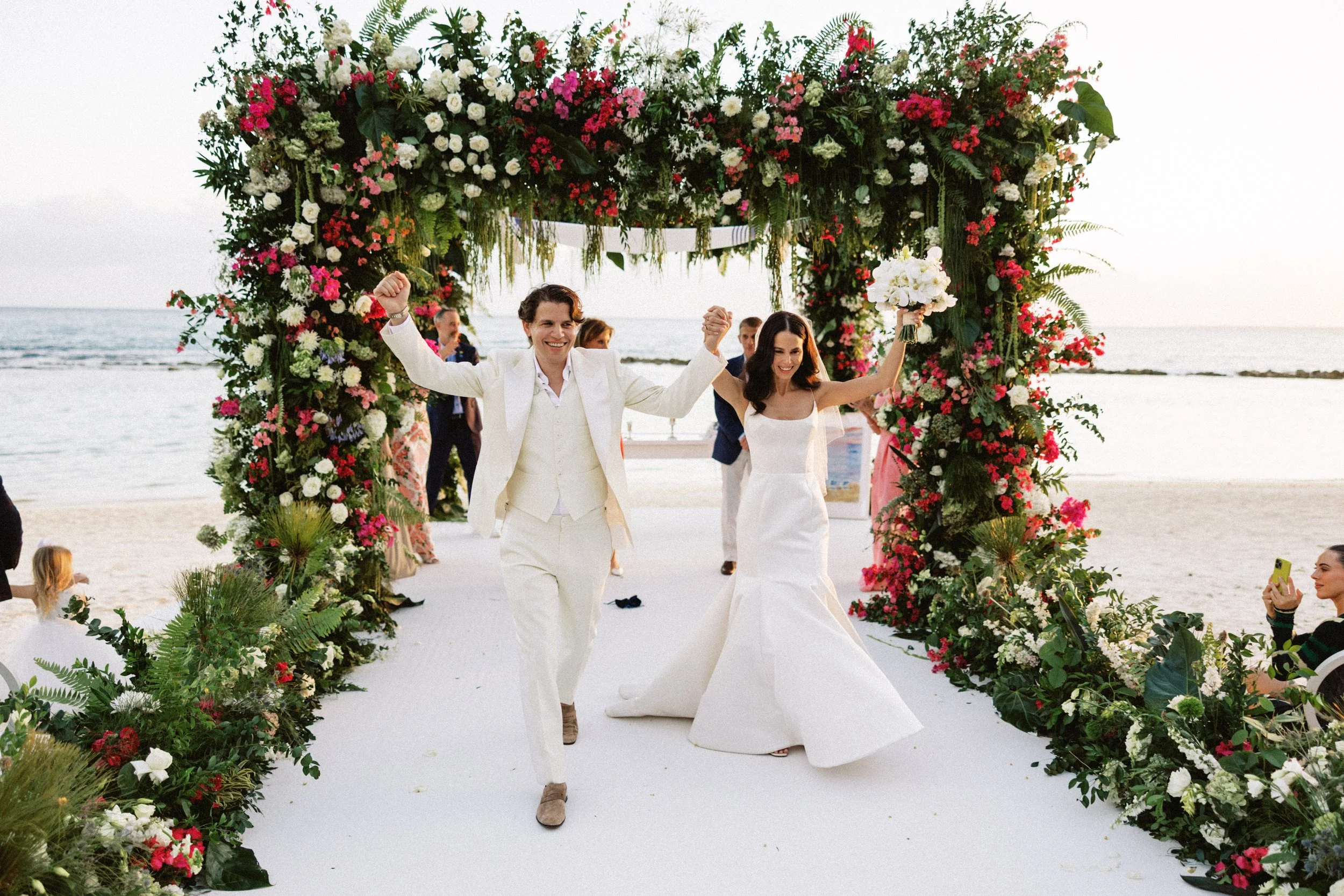 A newly married couple holding hands and smiling as they walk under a large floral arch on a beach, with friends and family in the background.