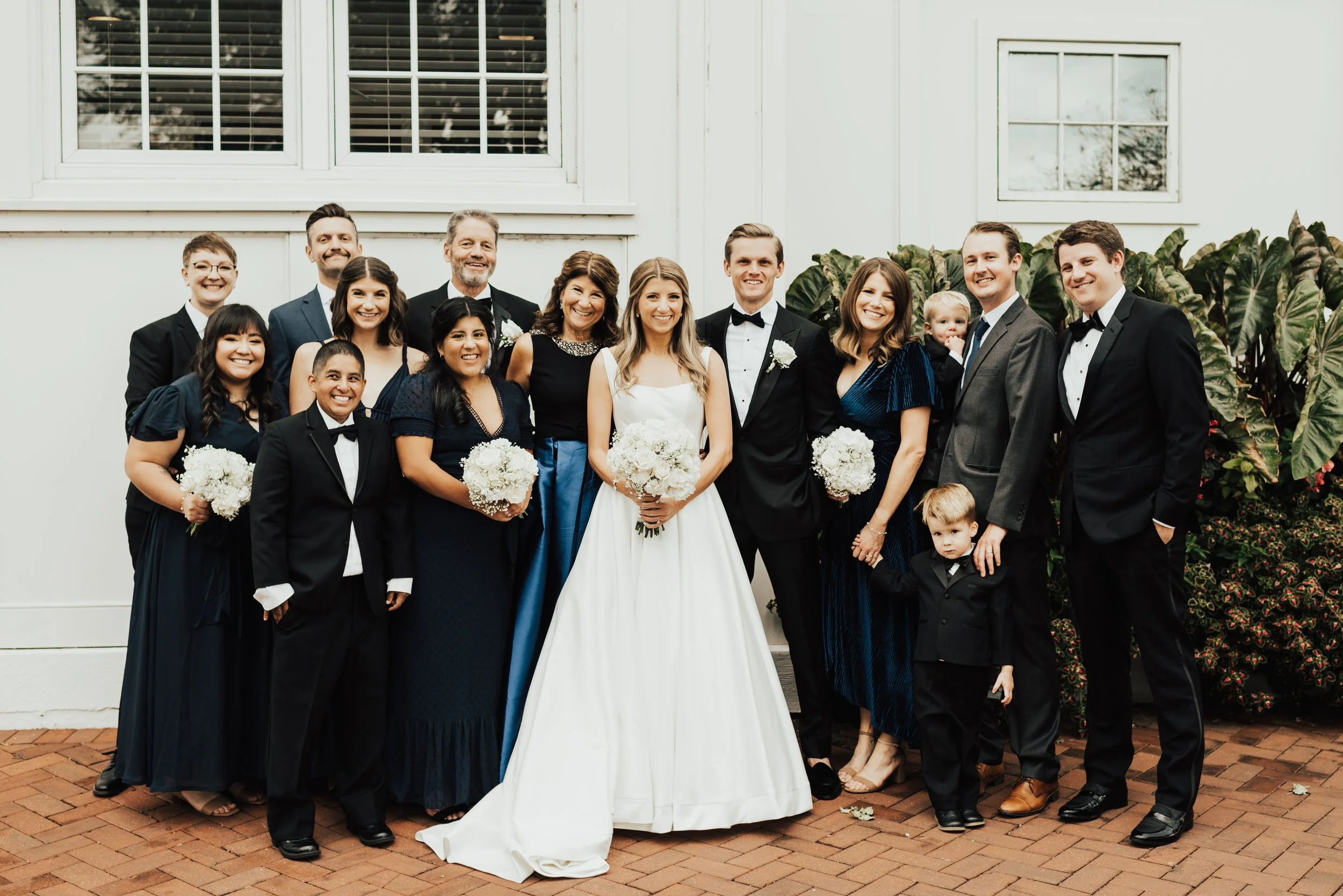 Group of people, including women in formal dresses and men in tuxedos, posing for a photo outside a house with white walls and windows, some holding bouquets of white flowers, during a celebration or event.