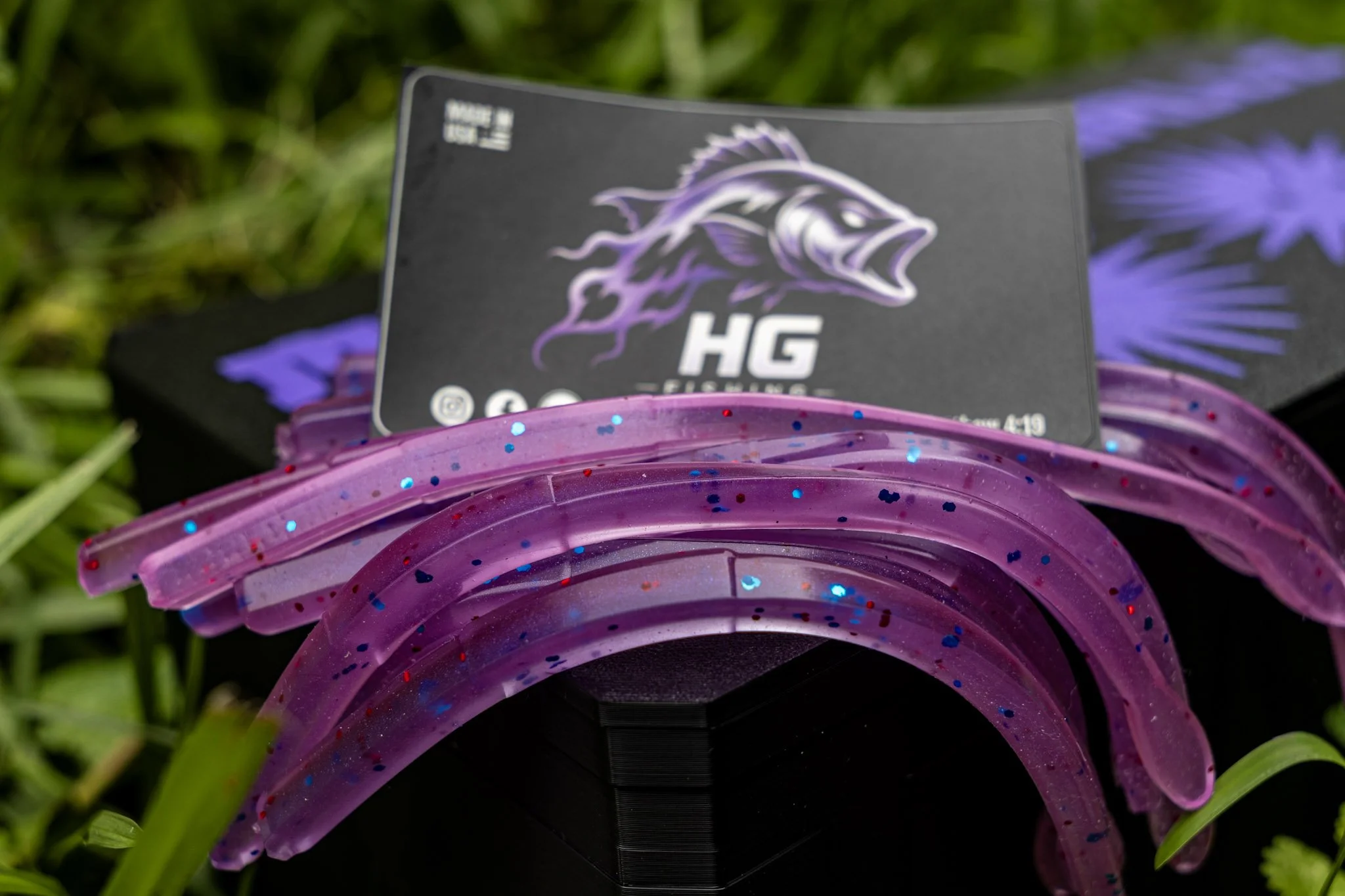 Close-up of a package of purple and glittery soft plastic fishing lures with a black card that has a logo of a purple fish and the text 'HG'. The lures are arranged in a fan shape on green grass.