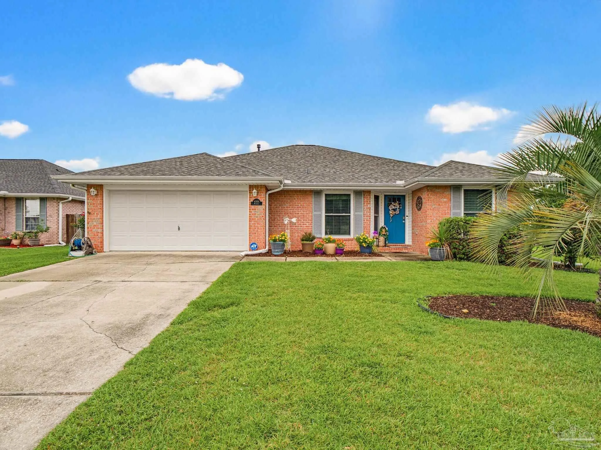 Front view of a single-story brick house with a two-car garage, a blue front door decorated with a wreath, potted plants and flowers in front, and a driveway with green lawn on either side. A palm tree is visible on the right side of the image, and a partly cloudy blue sky is above.