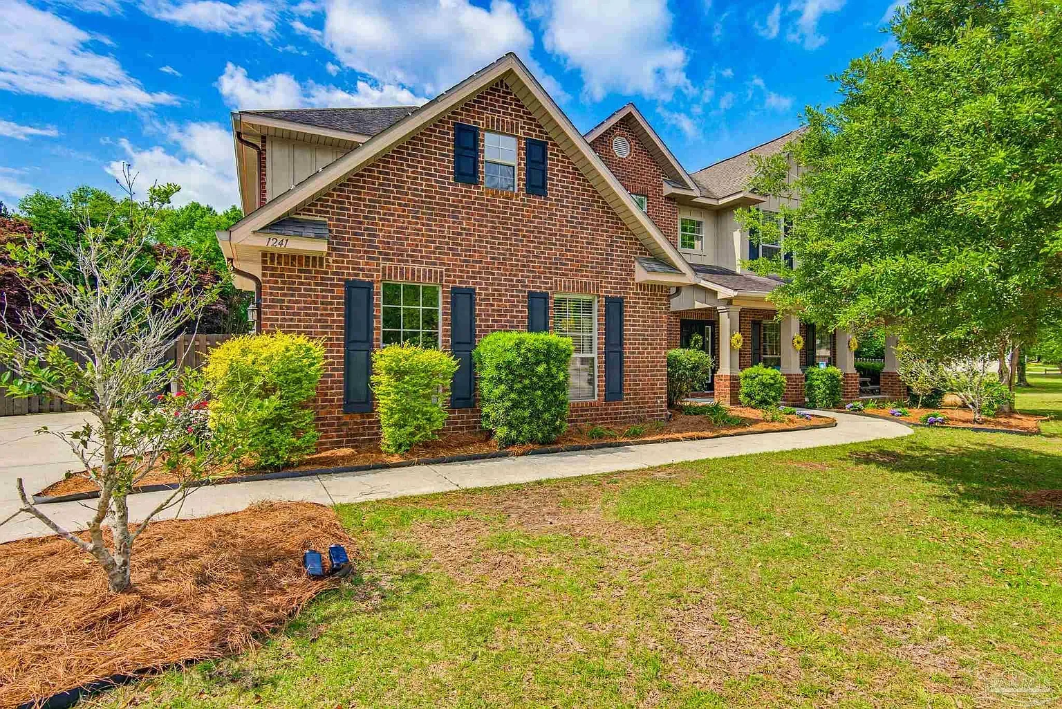 Front view of a two-story brick house with black shutters, a porch, and a neatly landscaped yard with bushes, trees, and a walkway under a blue sky with clouds.