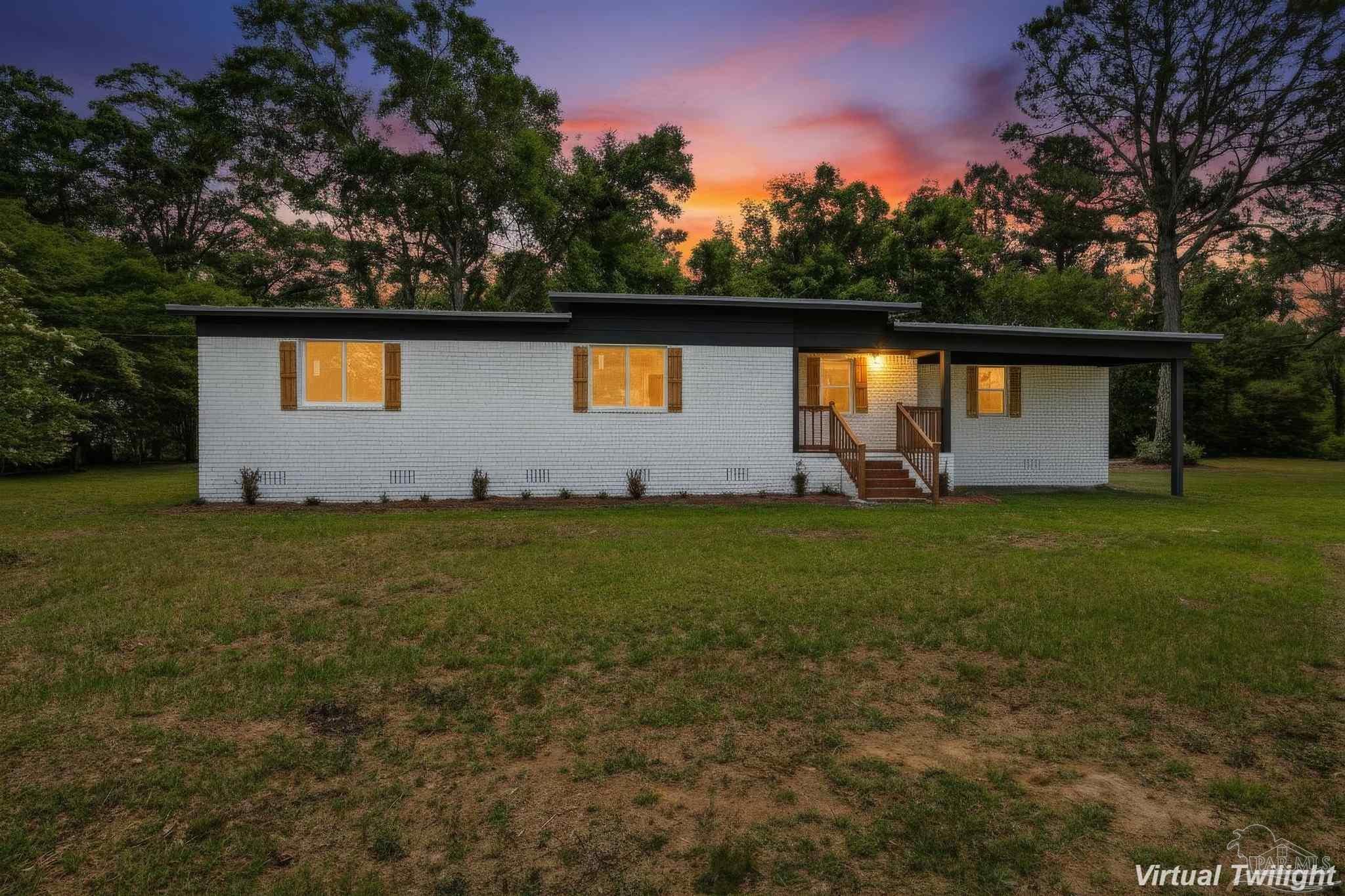 A small modern house with white brick walls, illuminated windows, and a small front porch with stairs, surrounded by green grass and trees under a colorful sunset sky.