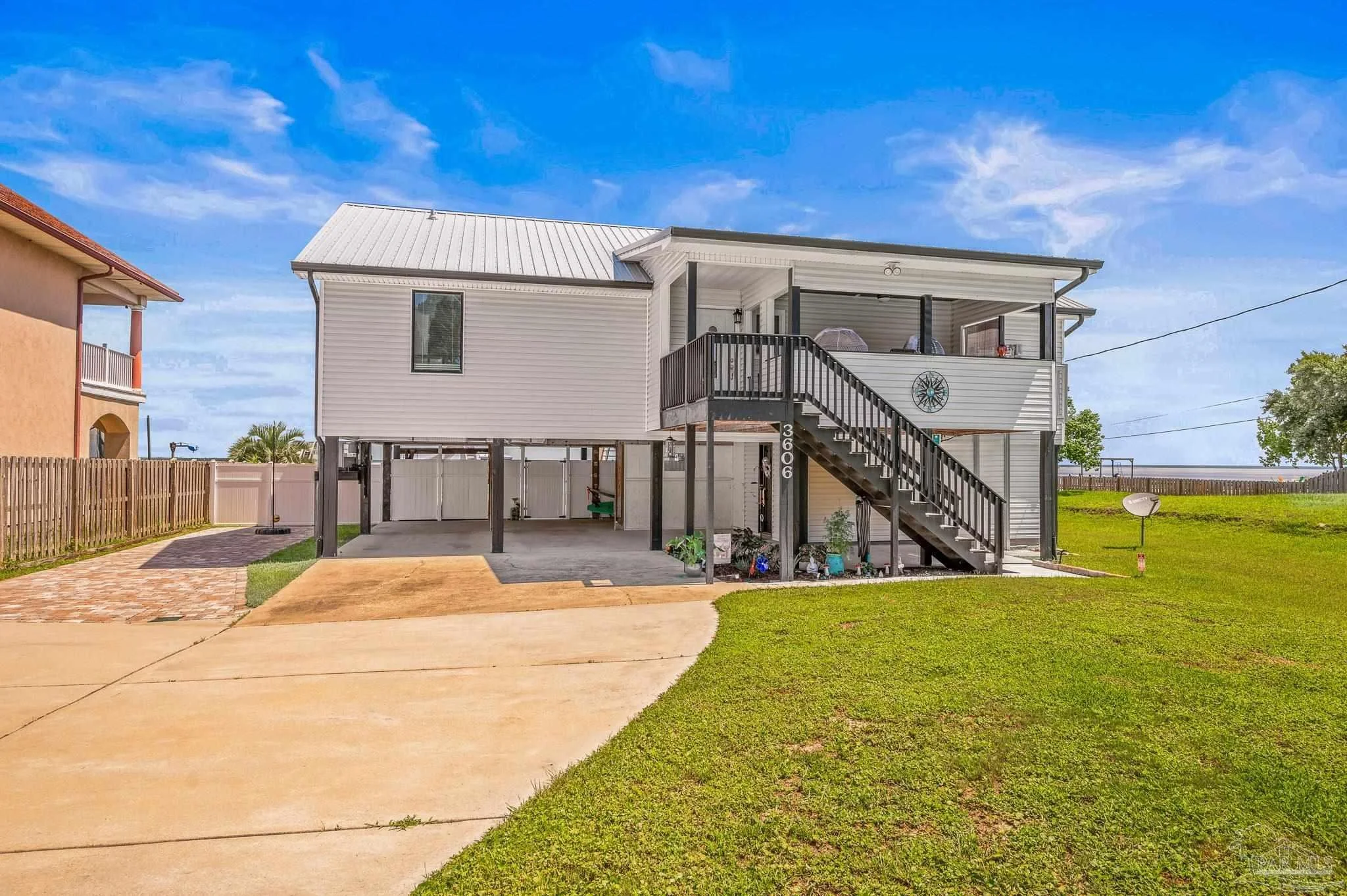 Backyard view of a two-story house with white siding and a screened-in porch on the second floor, wooden stairs, and a carport underneath. There is a concrete driveway, a fenced yard, and a well-maintained lawn with a clear blue sky.