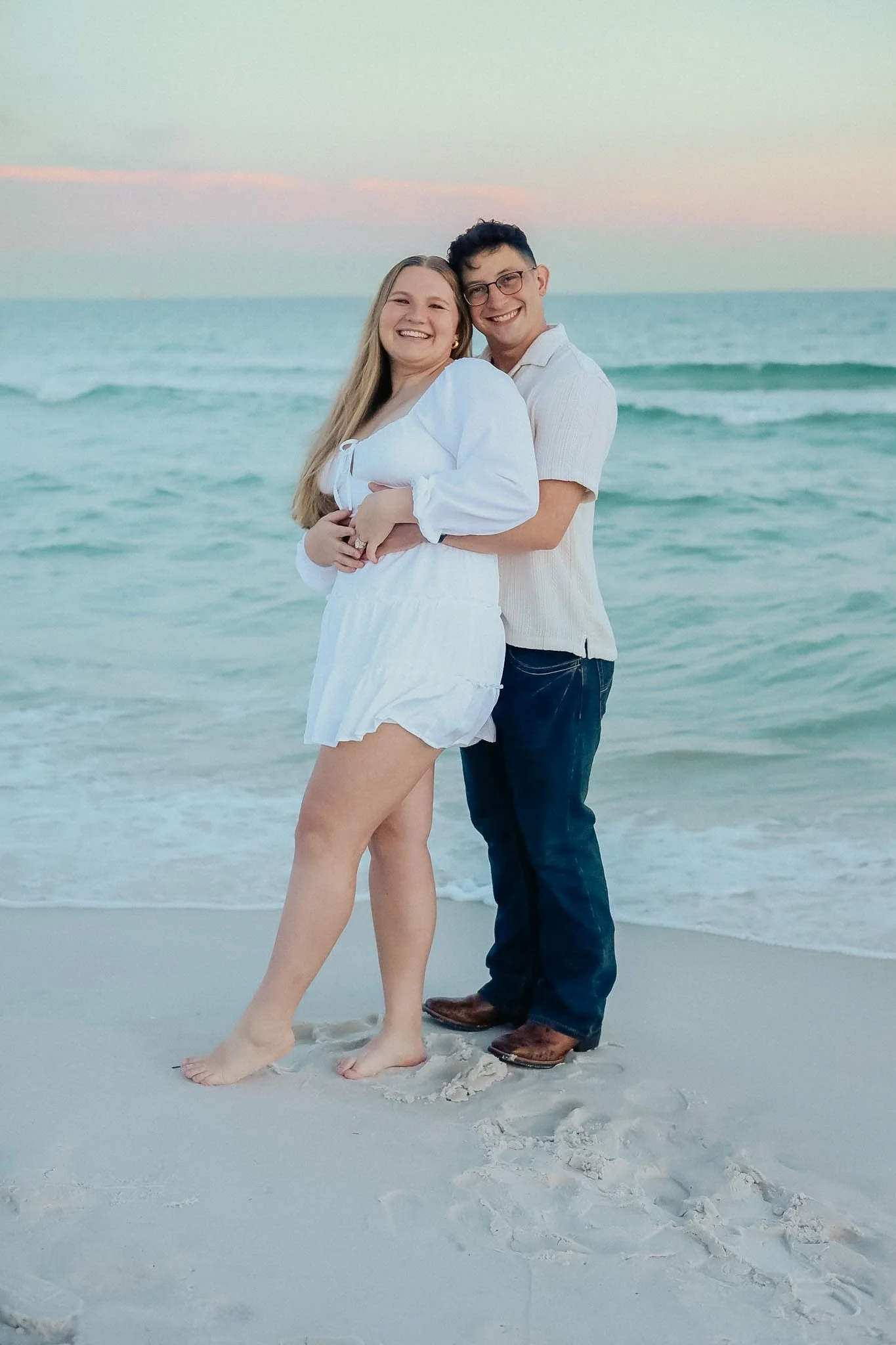 A smiling couple standing close together on a sandy beach, with the ocean and sunset sky in the background. The woman is wearing a white dress and the man is wearing a light-colored shirt and jeans.