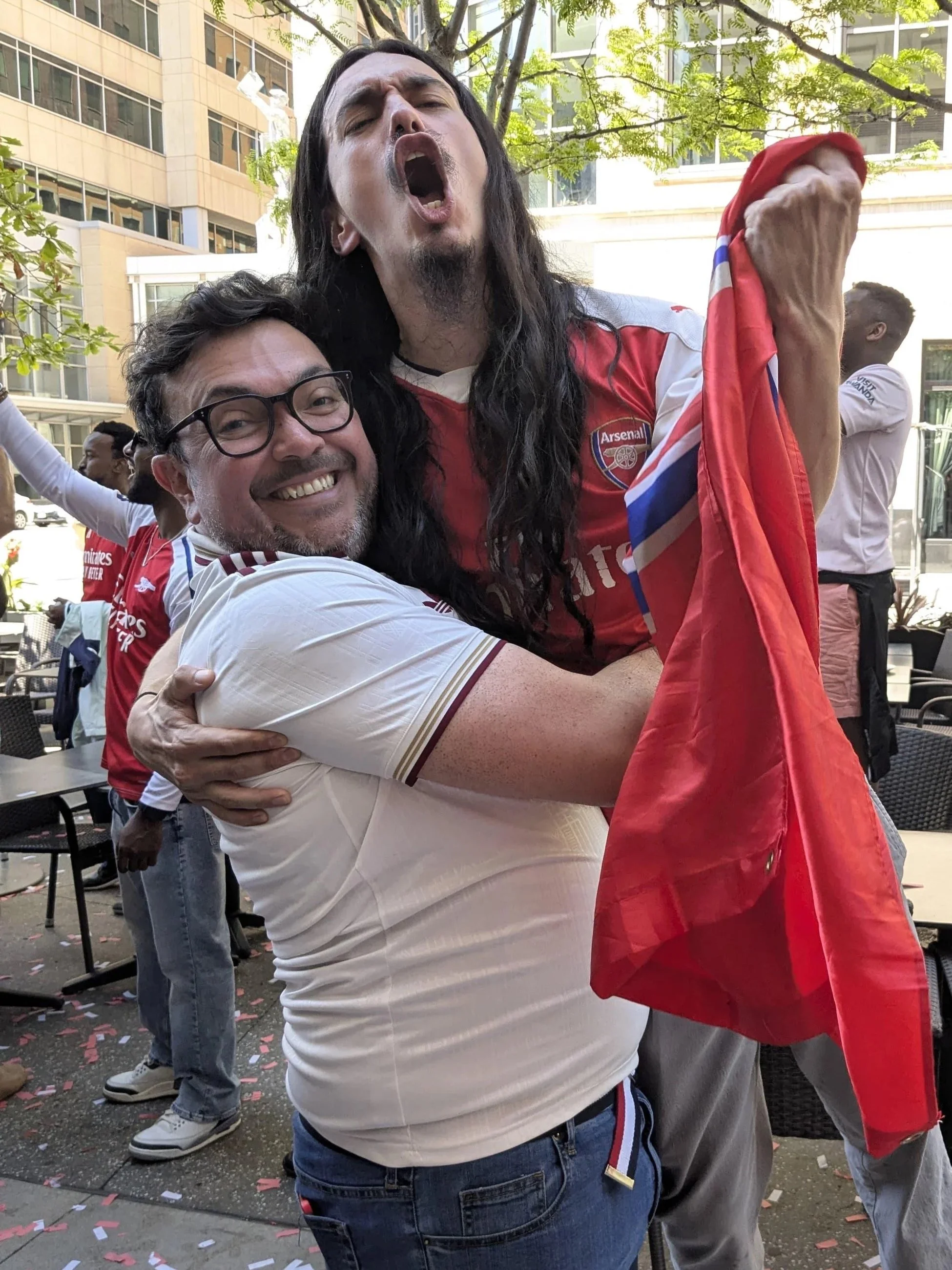 Two men celebrating outdoors, one wearing an Arsenal football jersey and holding a red scarf, the other smiling and hugging him. People and buildings are visible in the background.