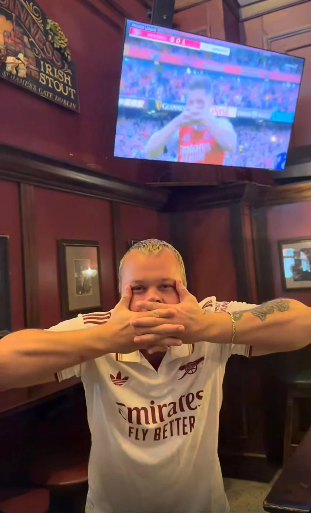 A man with platinum blonde hair and tattoos on his arms covering his mouth with his hands, sitting in a restaurant or pub with wood-paneled walls and framed pictures, wearing a white Arsenal football shirt that says 'miraTes Fly Better'. A television screen above shows a sports broadcast with a female athlete in a red and white uniform.