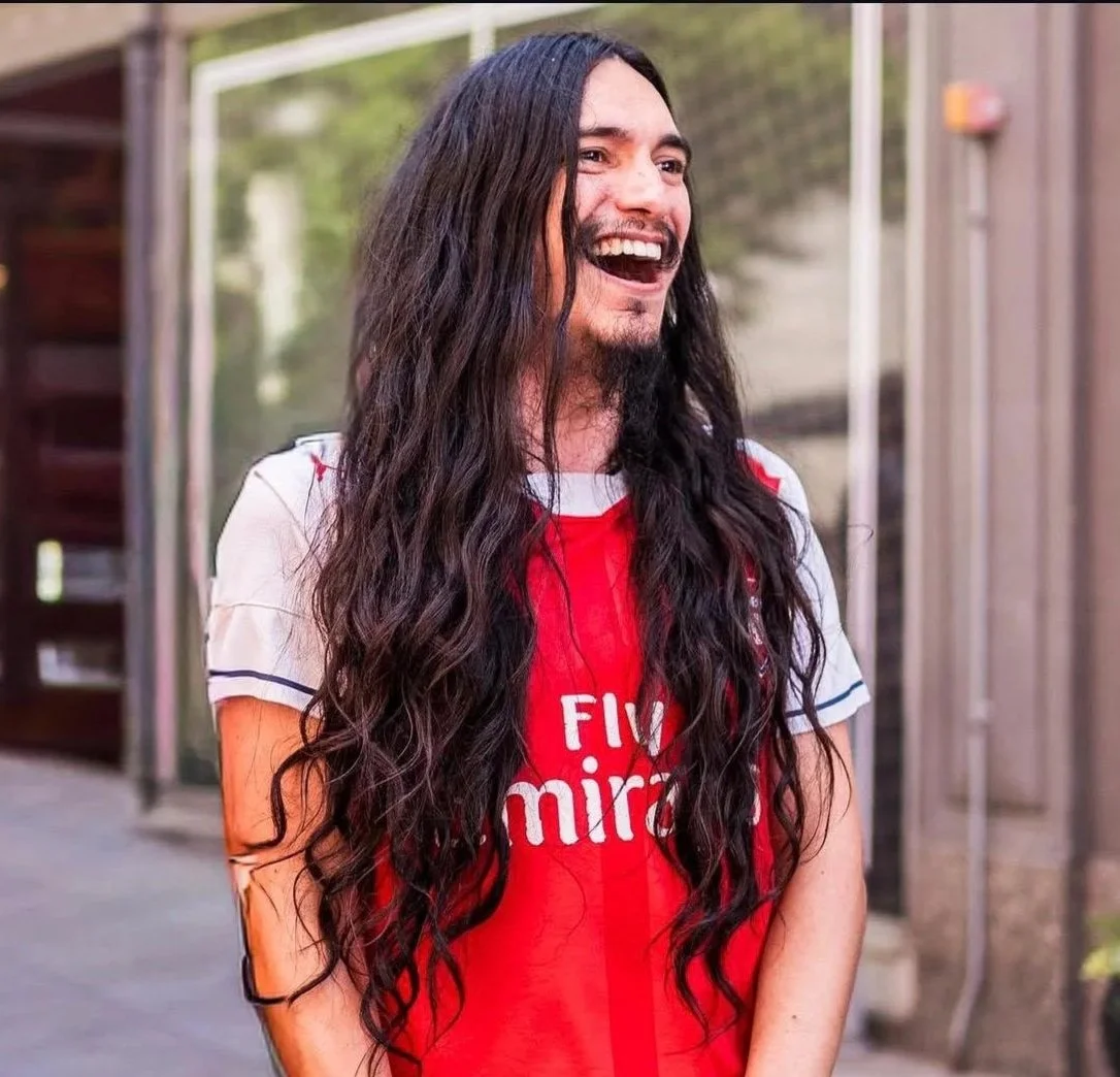 A man with long dark hair, smiling and wearing an Arsenal football jersey.