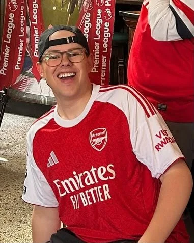 Young man smiling and wearing a red and white Arsenal soccer jersey with a black cap backwards at a sports event.