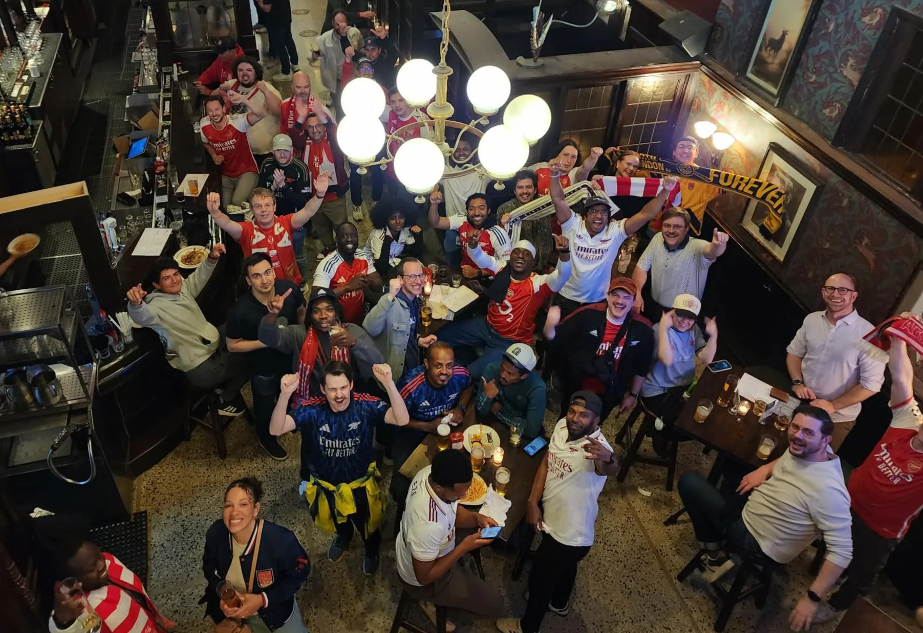A group of Arsenal soccer fans gathered in a bar celebrating, many wearing red and white jerseys, holding drinks, and cheering with raised fists, with some holding scarves and flags.