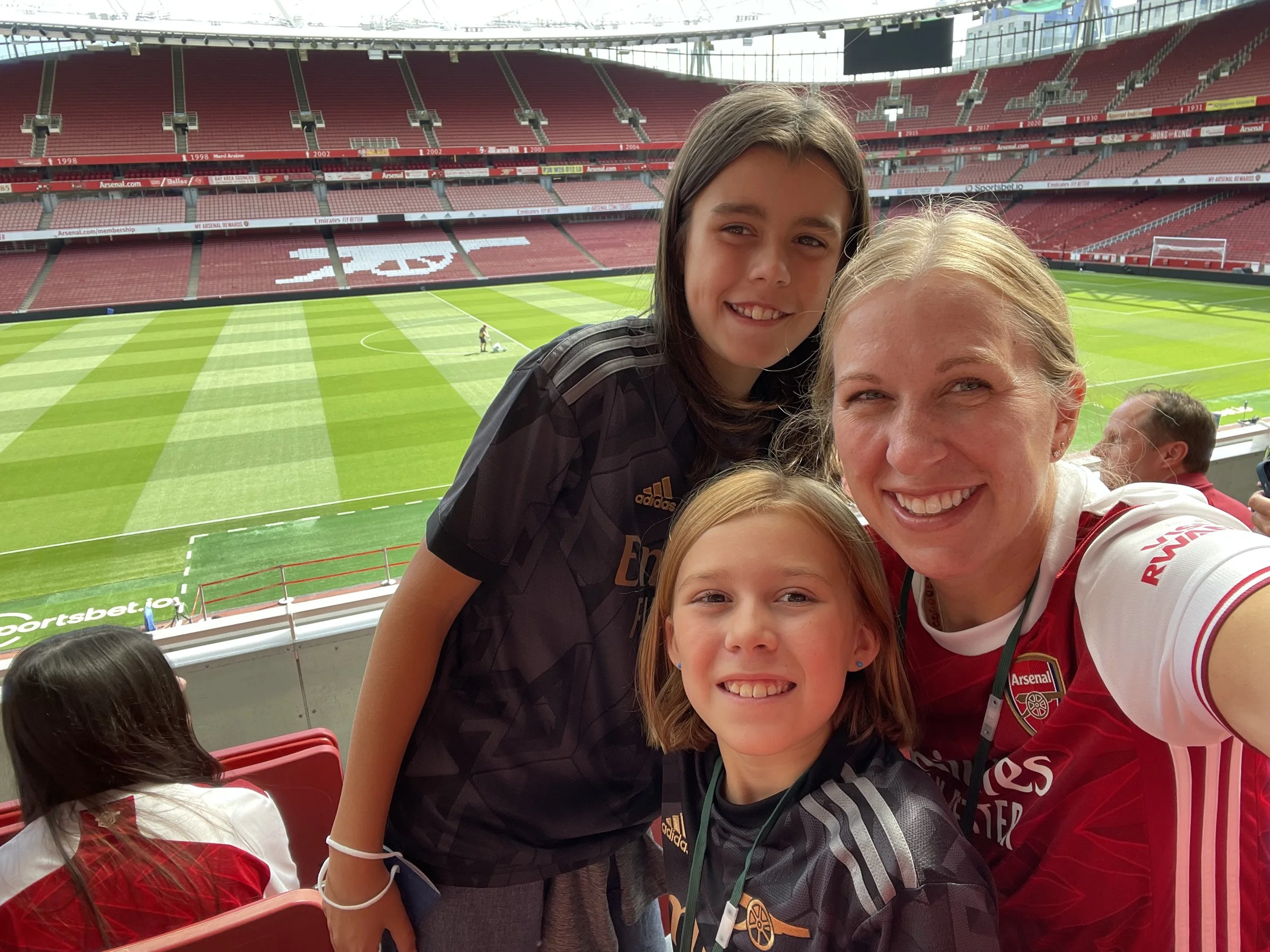 Group of smiling women and young girl taking selfie at Arsenal football stadium, with green grass field and empty red seats in background.