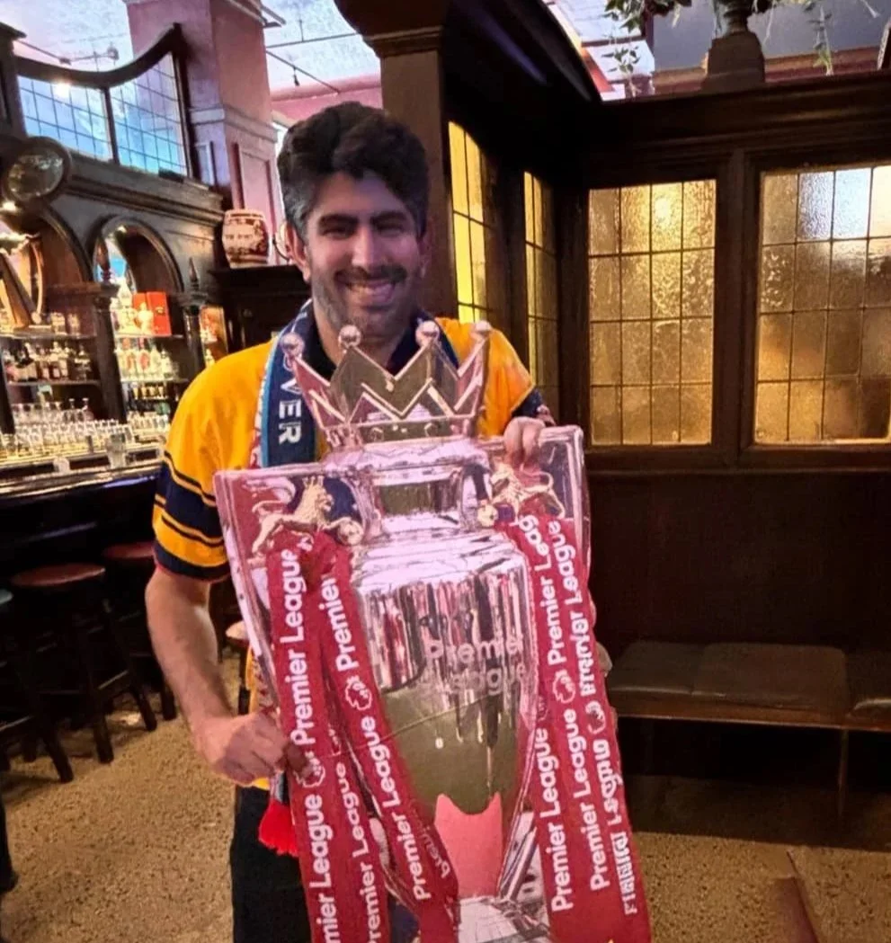 A man smiling and holding a large sign featuring a trophy and a crown, celebrating a sports victory in a pub or bar with wooden furniture and stained glass windows.