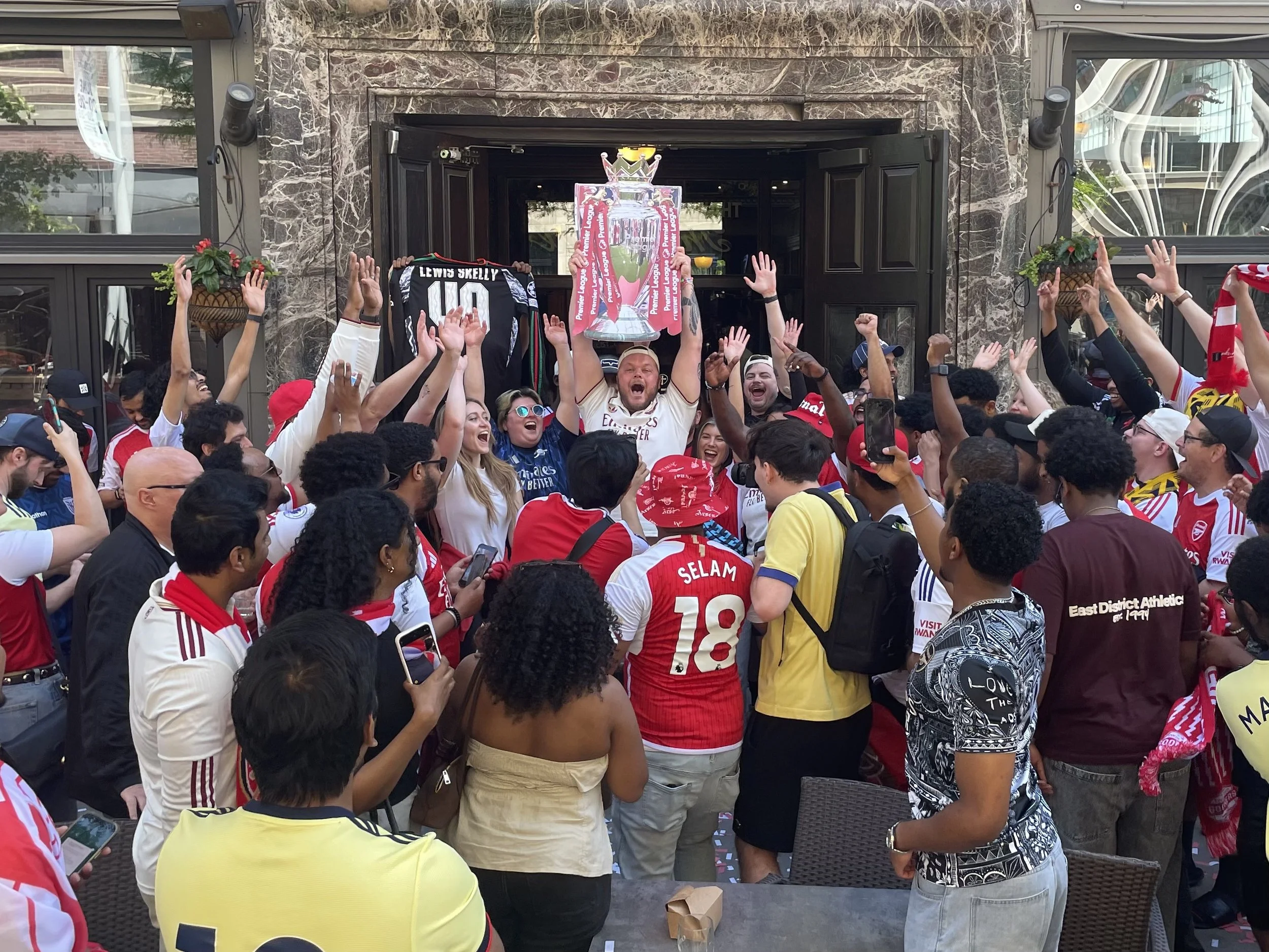A crowd of Arsenal soccer fans celebrating outside a building, holding a large trophy and jerseys, with many raising their hands in celebration.