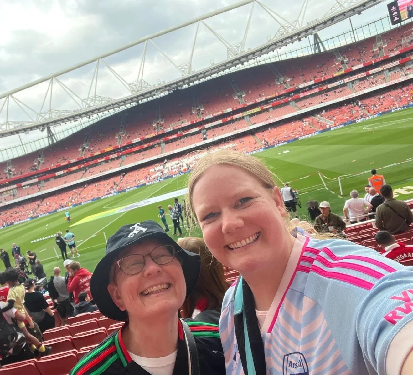 Two women smiling at a stadium, with empty red seats and a soccer field in the background. One woman is wearing a soccer jersey and the other has a hat and glasses.
