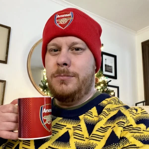 A man wearing an Arsenal beanie hat and a yellow and navy sweater, holding a red Arsenal mug, standing in front of a mirror with a Christmas tree in the background.