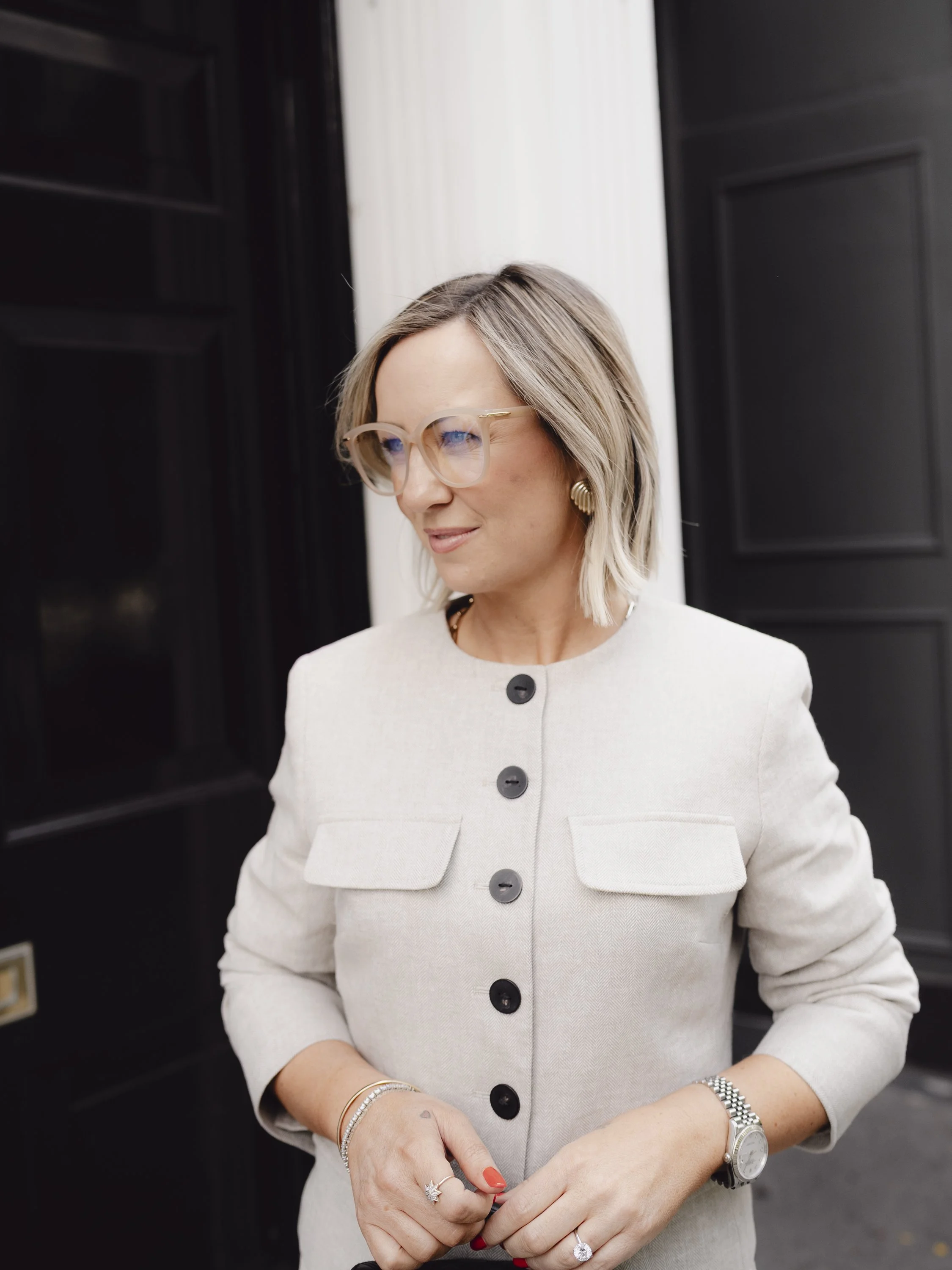 A woman with blonde hair styled in a bob, wearing glasses, gold earrings, and a light beige jacket with black buttons, standing outdoors near black doors or cabinets.