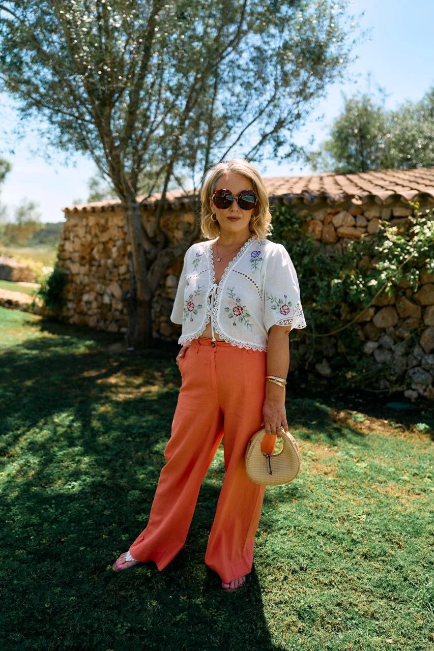 Woman with blonde hair wearing large sunglasses, a white embroidered blouse, orange wide-leg pants, holding a small beige handbag, standing on grass with trees and a stone wall in the background.