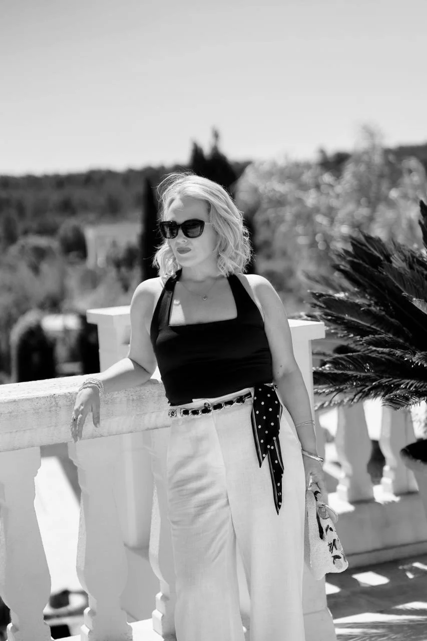 A woman with blonde hair wearing sunglasses, a black tank top, and white high-waisted pants, standing outdoors by a white railing with trees and sky in the background.