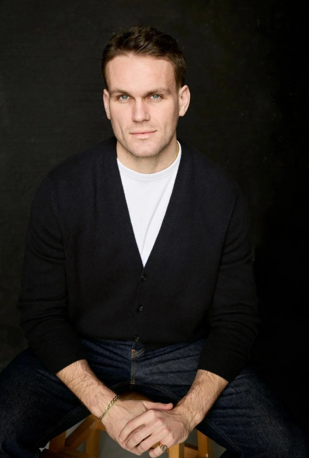 A young man with short brown hair and blue eyes, wearing a white shirt, black cardigan, and dark jeans, sitting on a wooden stool against a black background.