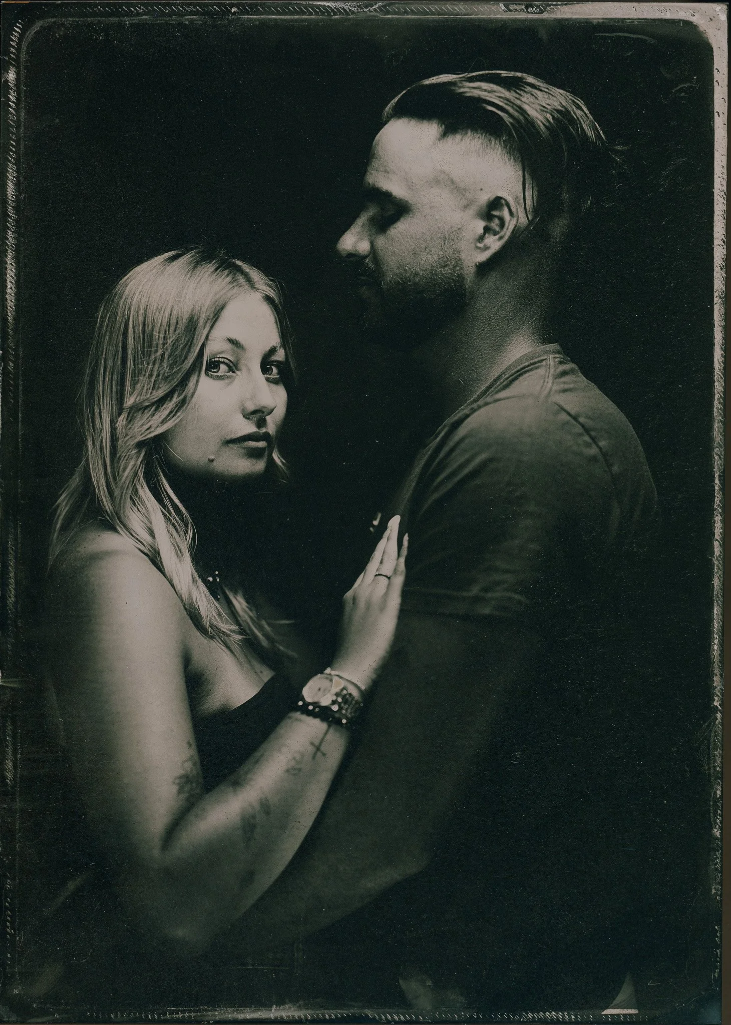 Une femme blonde portant un vêtement noir, avec plusieurs tatouages, regarde d'un côté, avec une main touchant la poitrine d'un homme chauve, barbu. L'homme est vu de profil, avec des cheveux longs attachés à l'arrière. La photo est en noir et blanc.