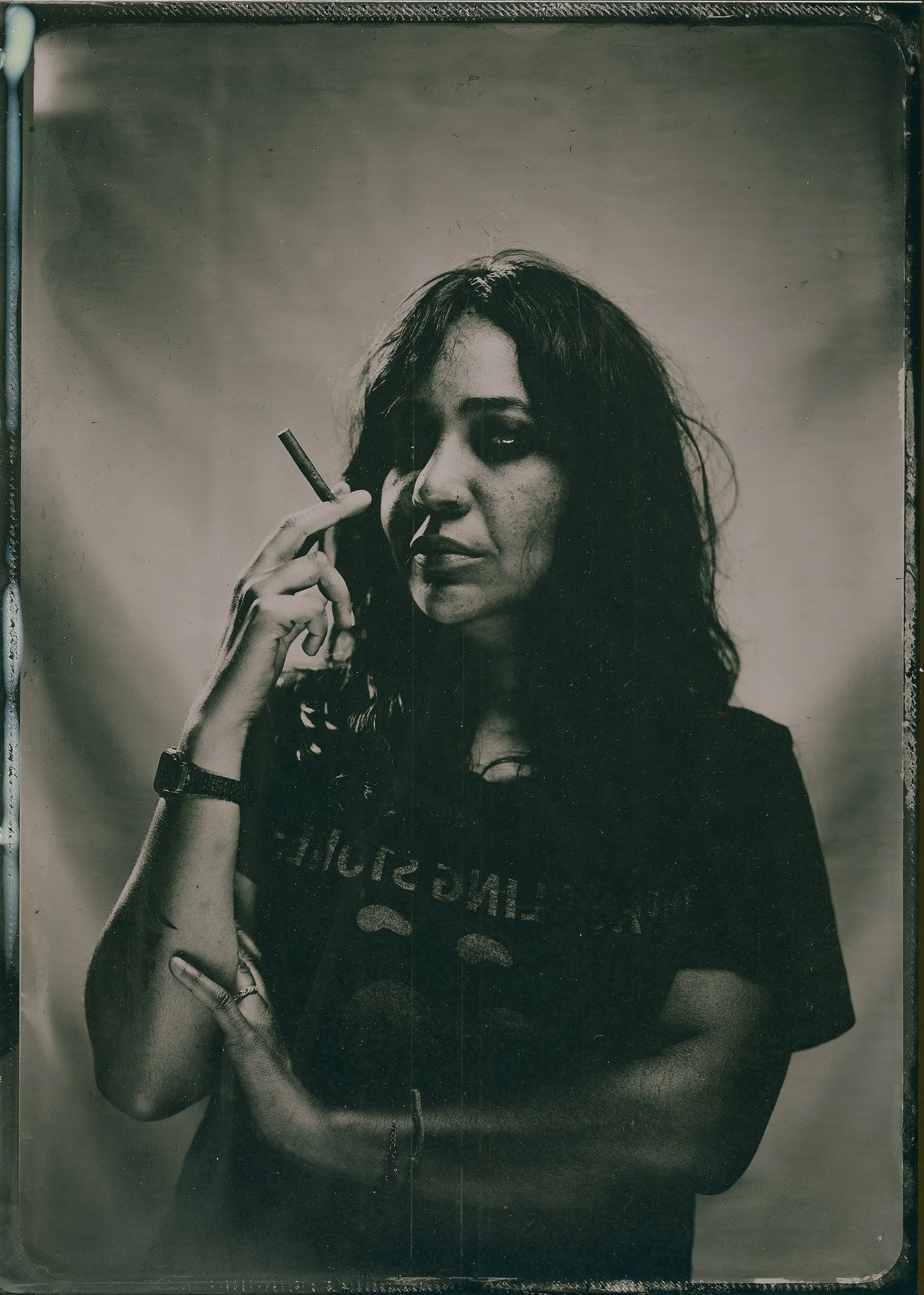 Photo en noir et blanc d'une femme avec de longs cheveux, portant un bracelet à la montre, tenant un cigarette à la main, et portant un t-shirt avec un logo partiellement visible.