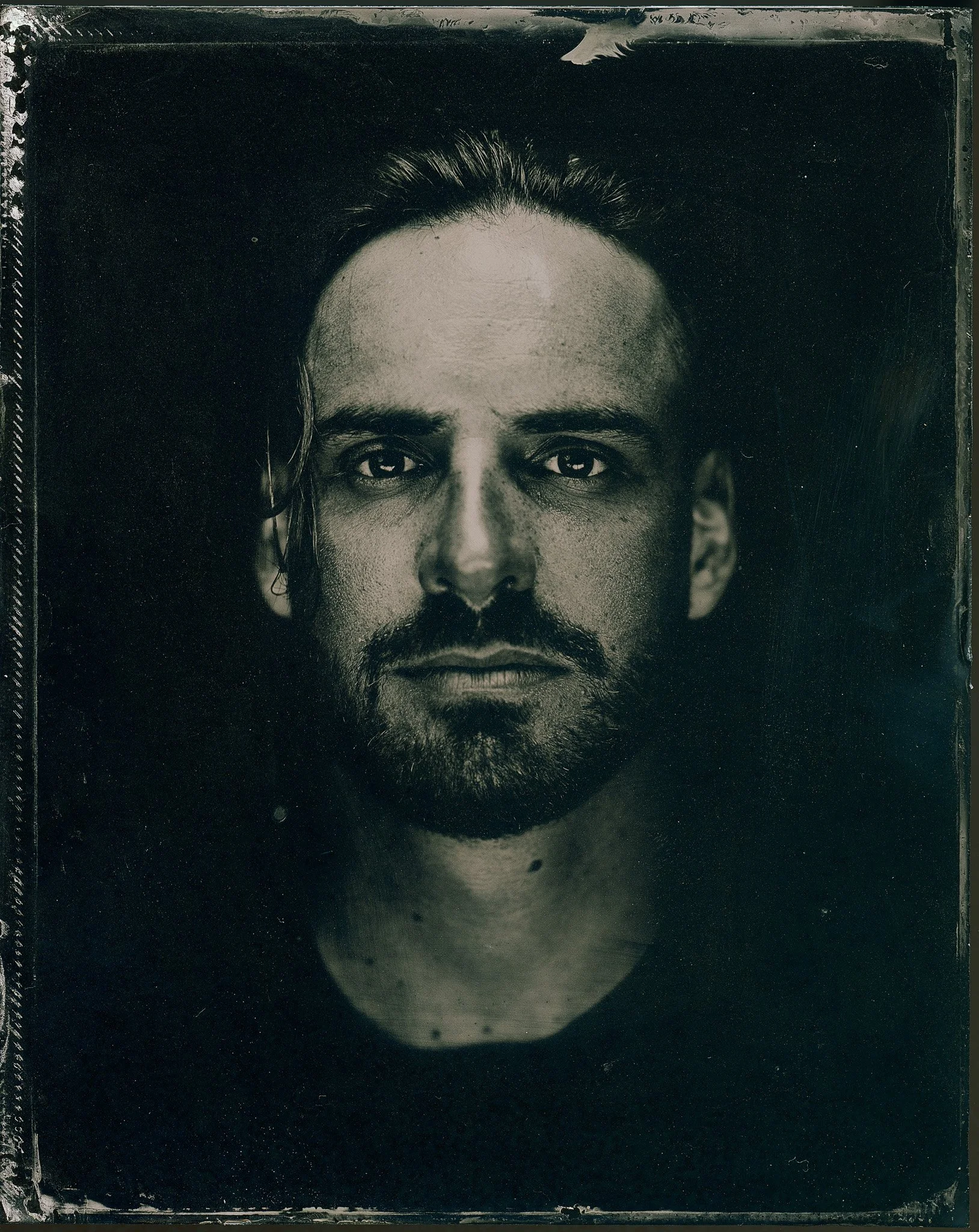 Portrait en noir et blanc d'une personne avec un visage pleinement visible, fond noir, une expression neutre, cheveux longs et facial hair.