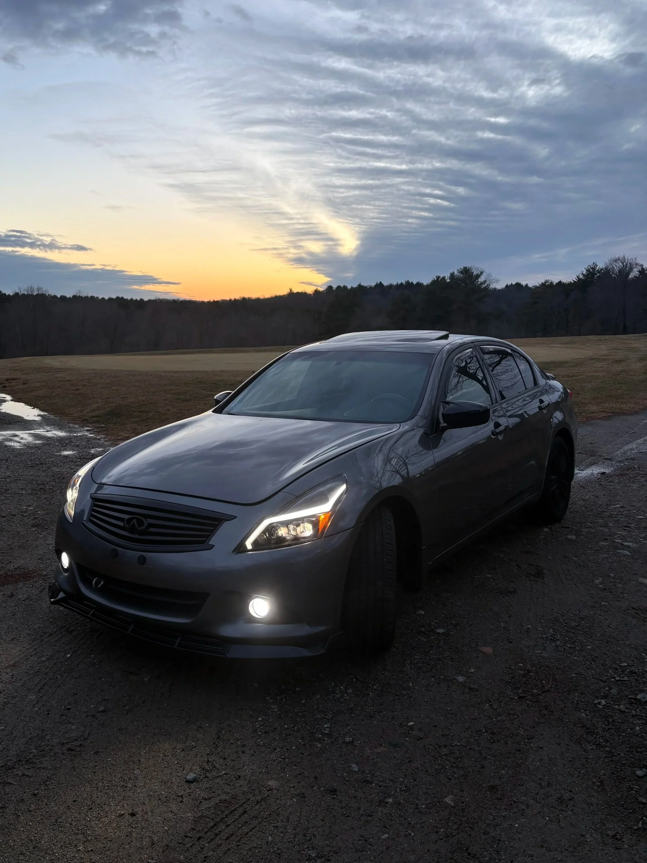 A gray Infiniti sedan parked on a dirt surface with a scenic landscape of trees and a sunset sky in the background.