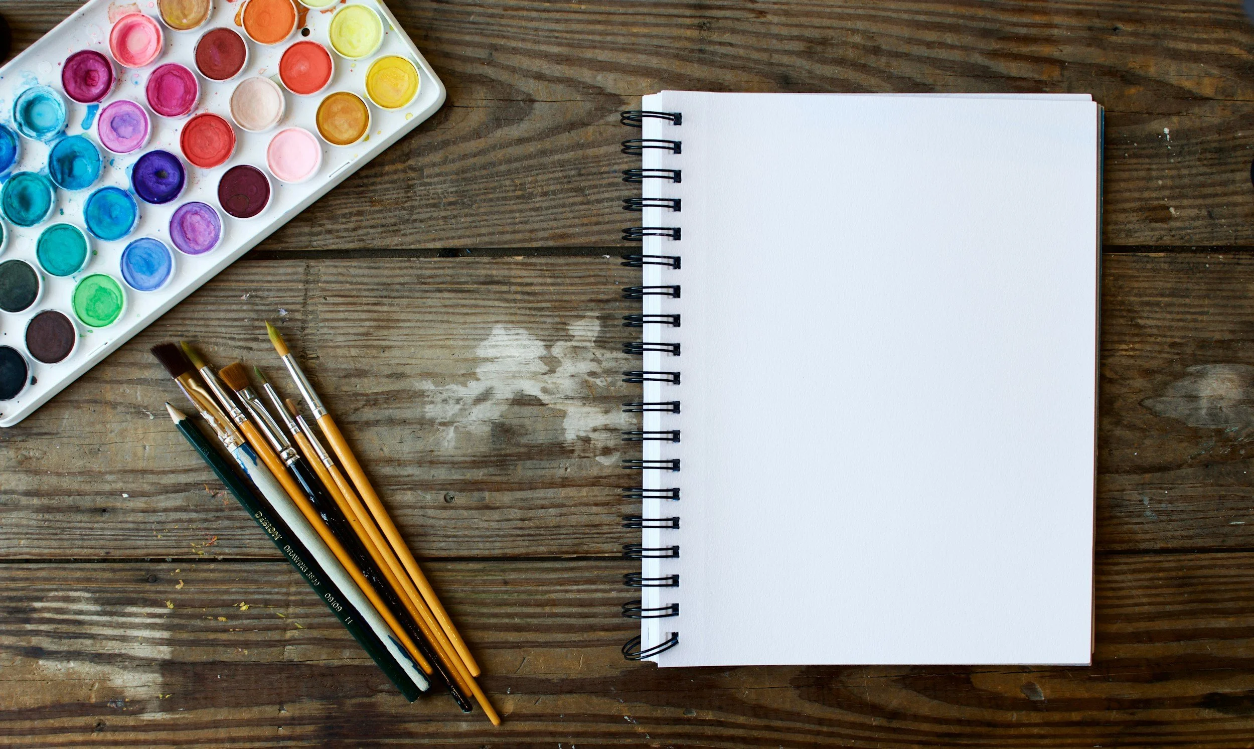 A watercolor paint set, paint brushes, and a blank spiral notebook on a wooden surface.