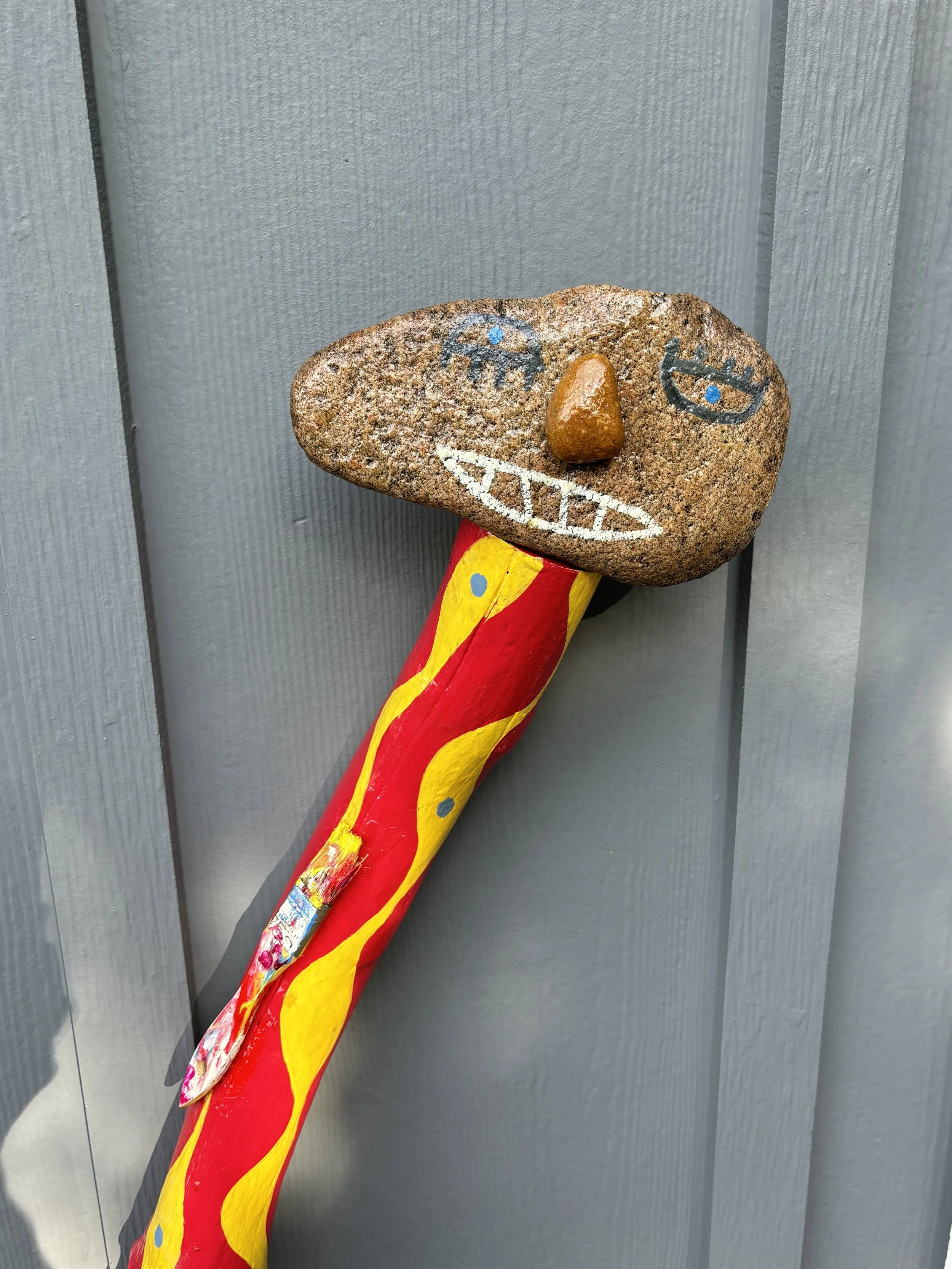 A rock with a painted face, including eyes, a wide smile, and a nose, is attached to a painted wooden stick with red and yellow stripes. The background is a gray vertical panel wall.