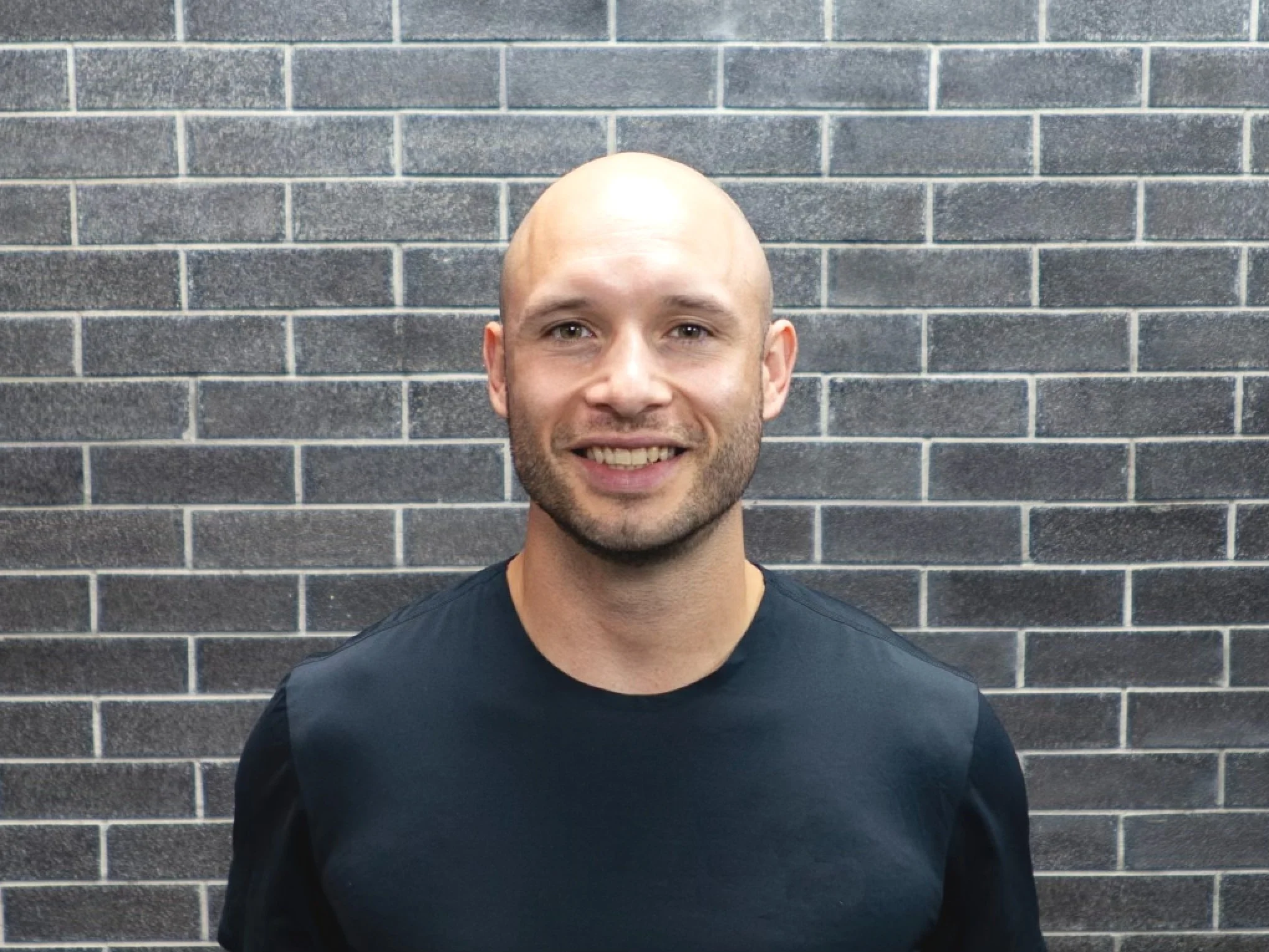 Portrait of a smiling bald man with facial hair, wearing a black shirt, standing in front of a gray brick wall.