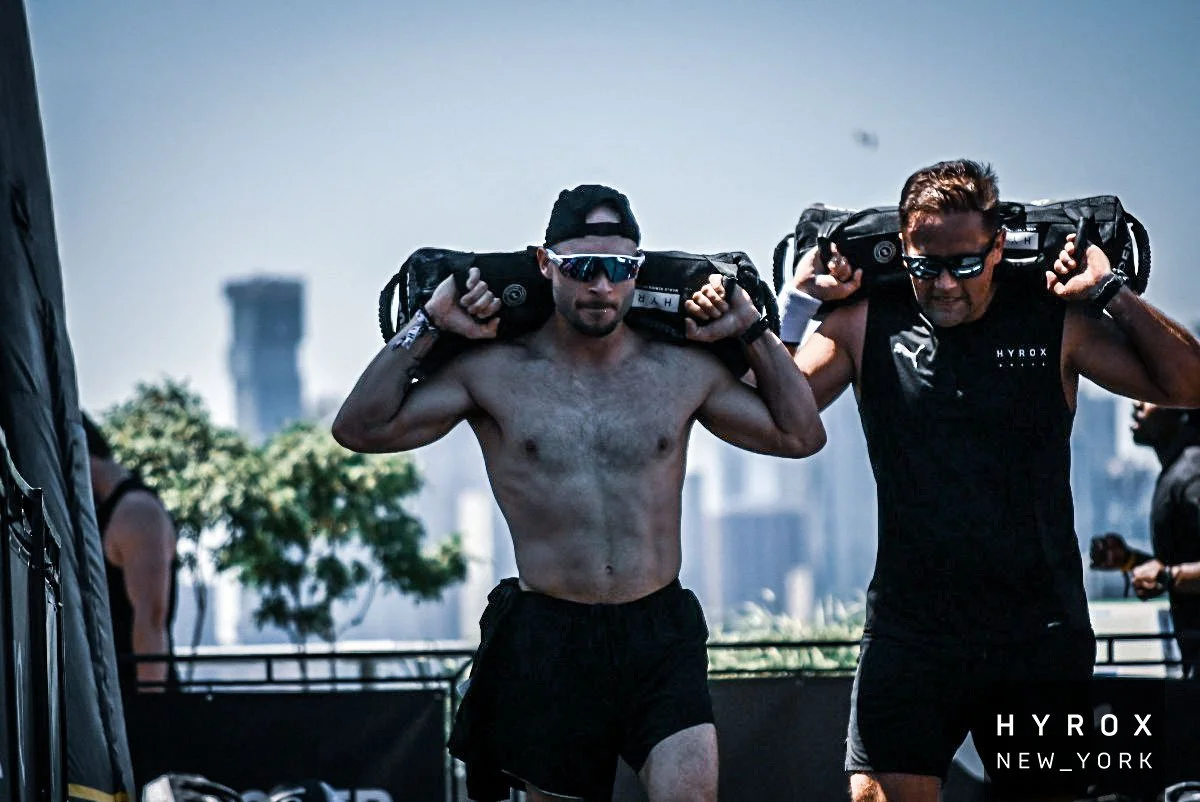 Two men carrying large backpacks on their shoulders during a fitness event outdoors, with a city skyline in the background and the logo 'HYROX NEW_YORK' in the bottom right corner.