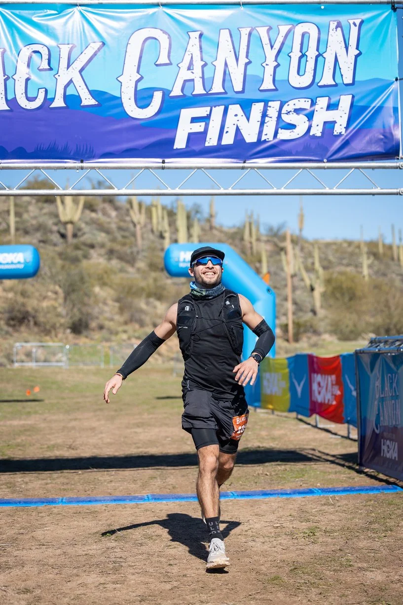 Male runner in black athletic gear finishing the Black Canyon race, smiling as he crosses the finish line with a large blue banner overhead that says "Black Canyon Finish" and desert landscape with cacti in the background.