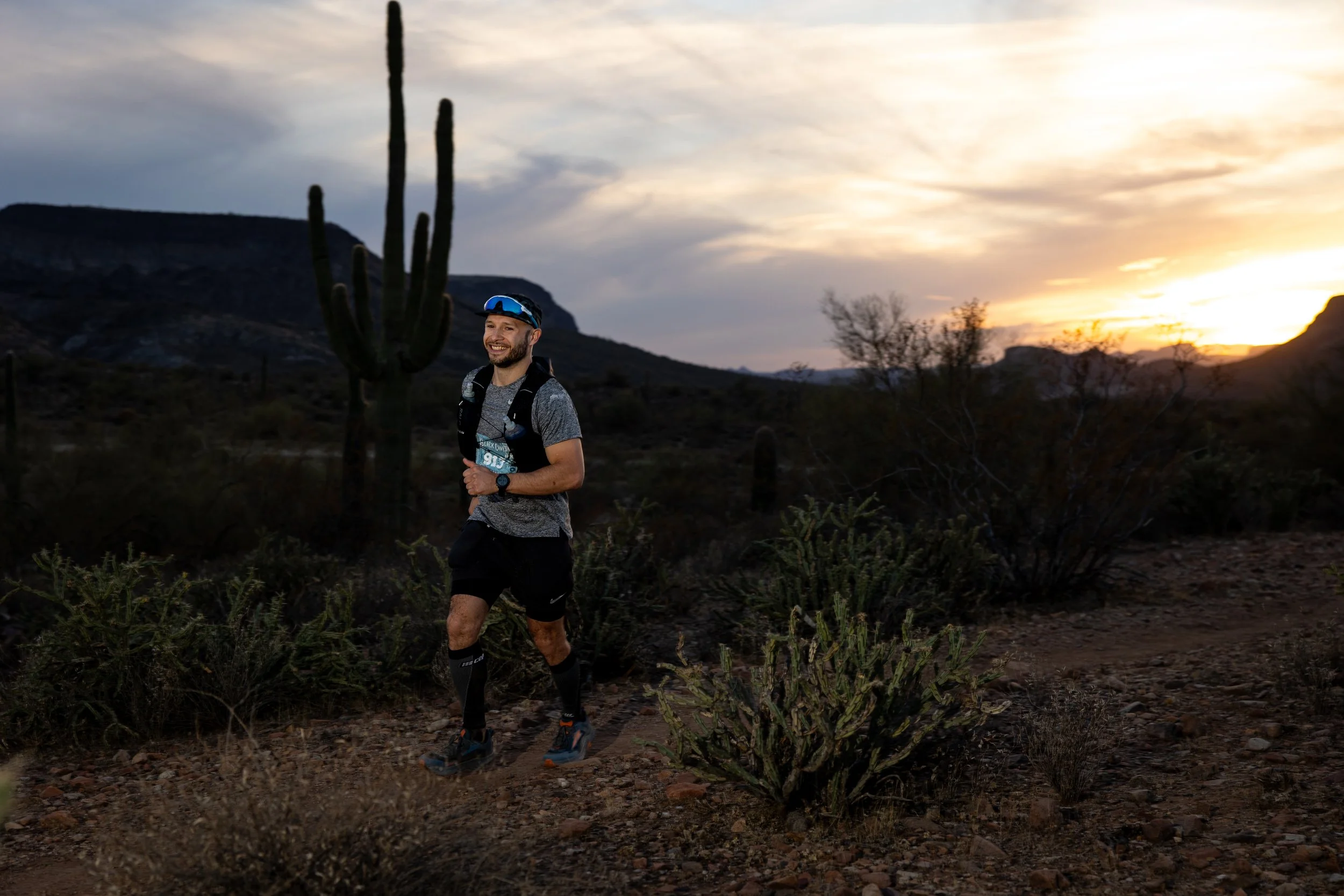 A man trail running in a desert landscape at sunset, wearing a gray shirt, black shorts, and a hydration pack, with cacti and distant mountains in the background.