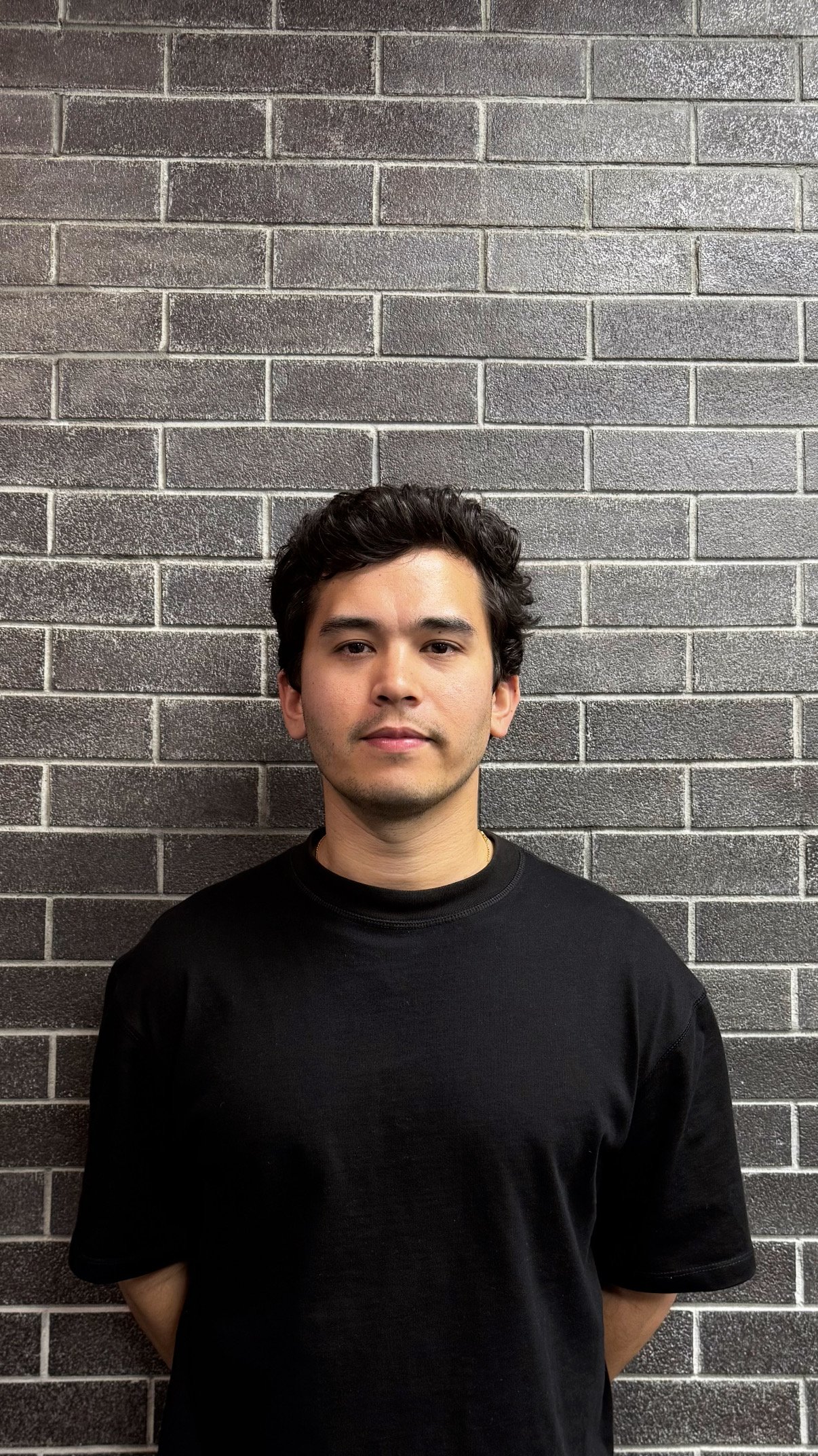 A young man with dark, curly hair and light skin is standing against a gray brick wall. He is wearing a plain black T-shirt and looks directly at the camera with a neutral expression.