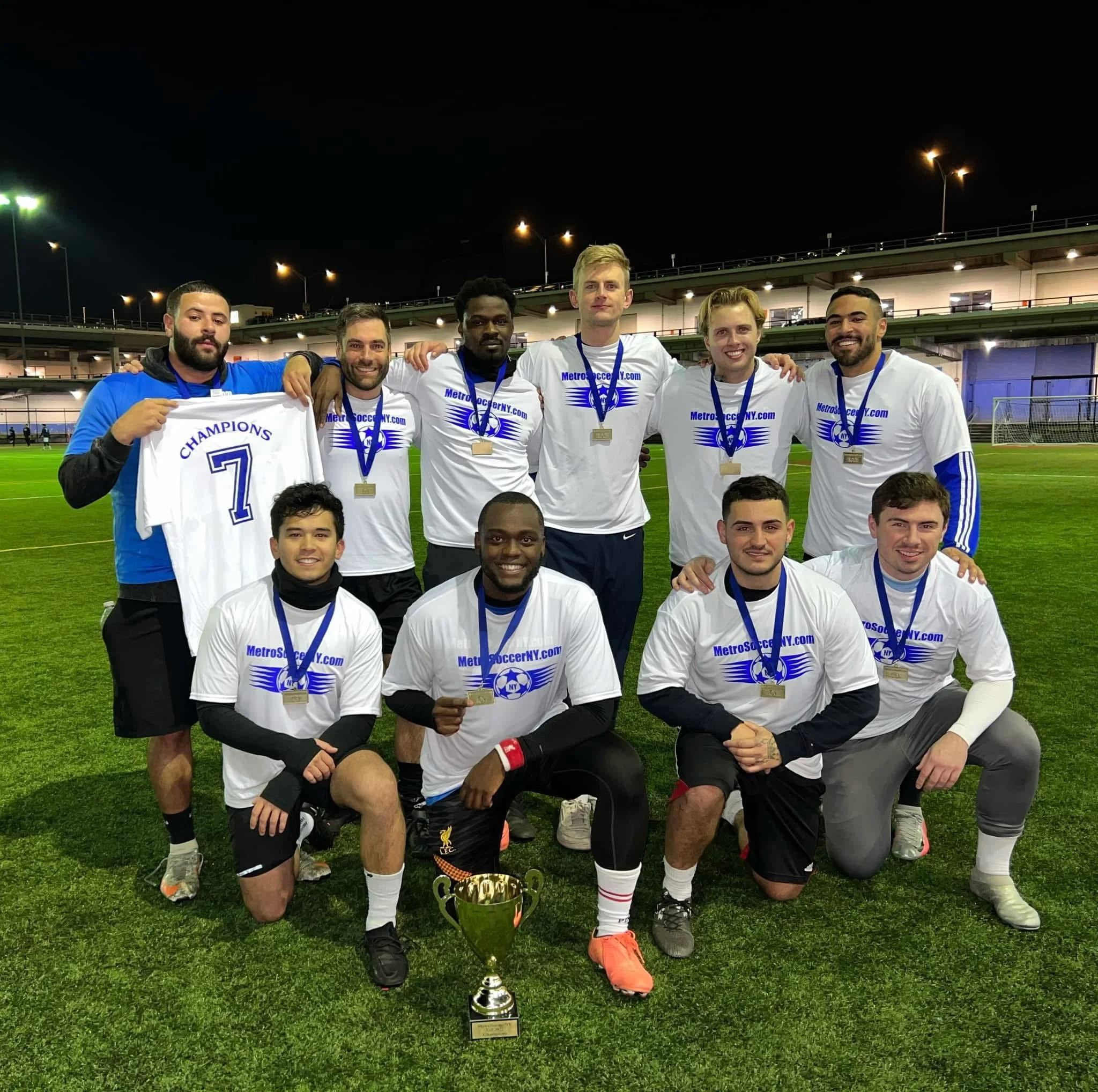 A soccer team celebrating on the field with medals and a trophy after winning a championship, with players kneeling and standing together at night under stadium lights.