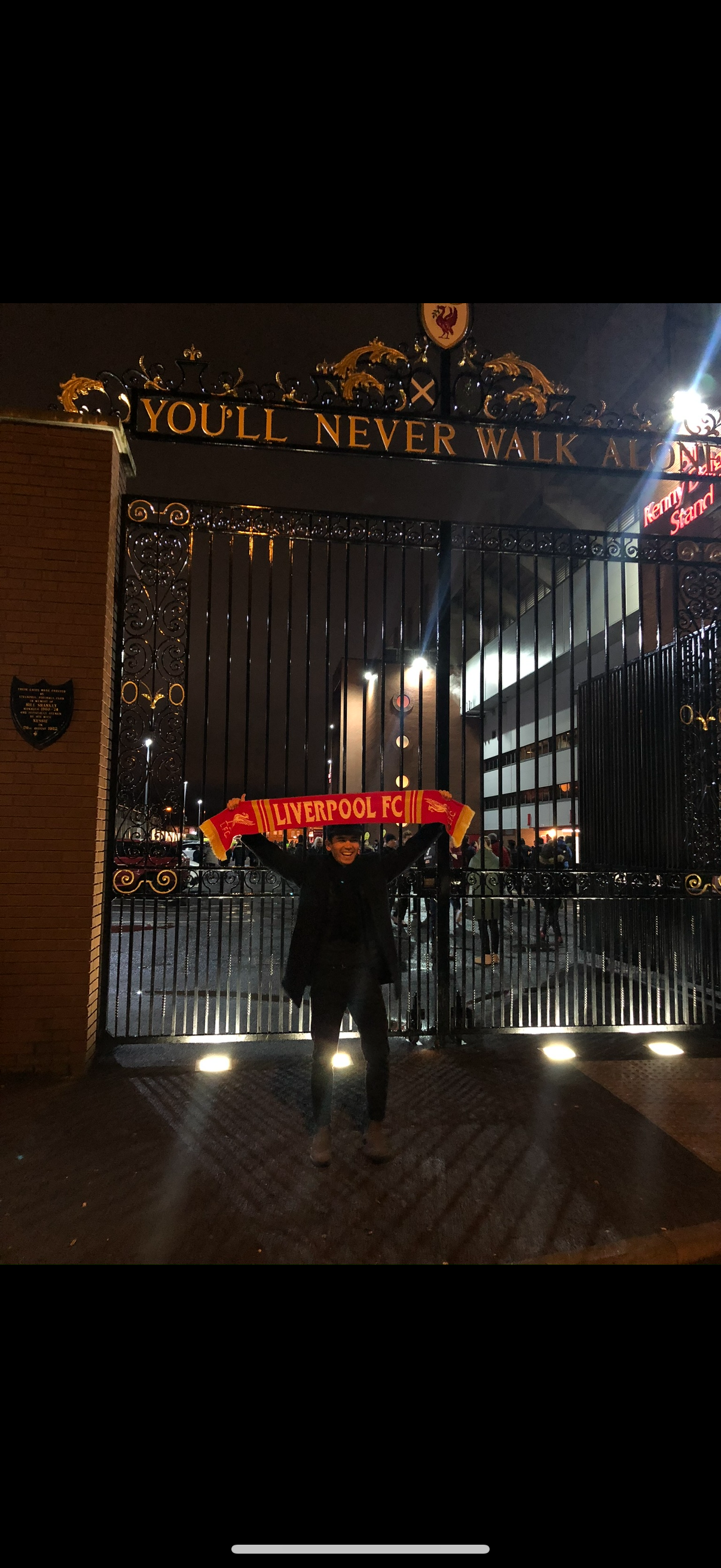 A person standing in front of a gated entrance holding a red Liverpool FC scarf. The scene is at night with streetlights and a sign above the gate that reads 'You'll Never Walk Alone'.