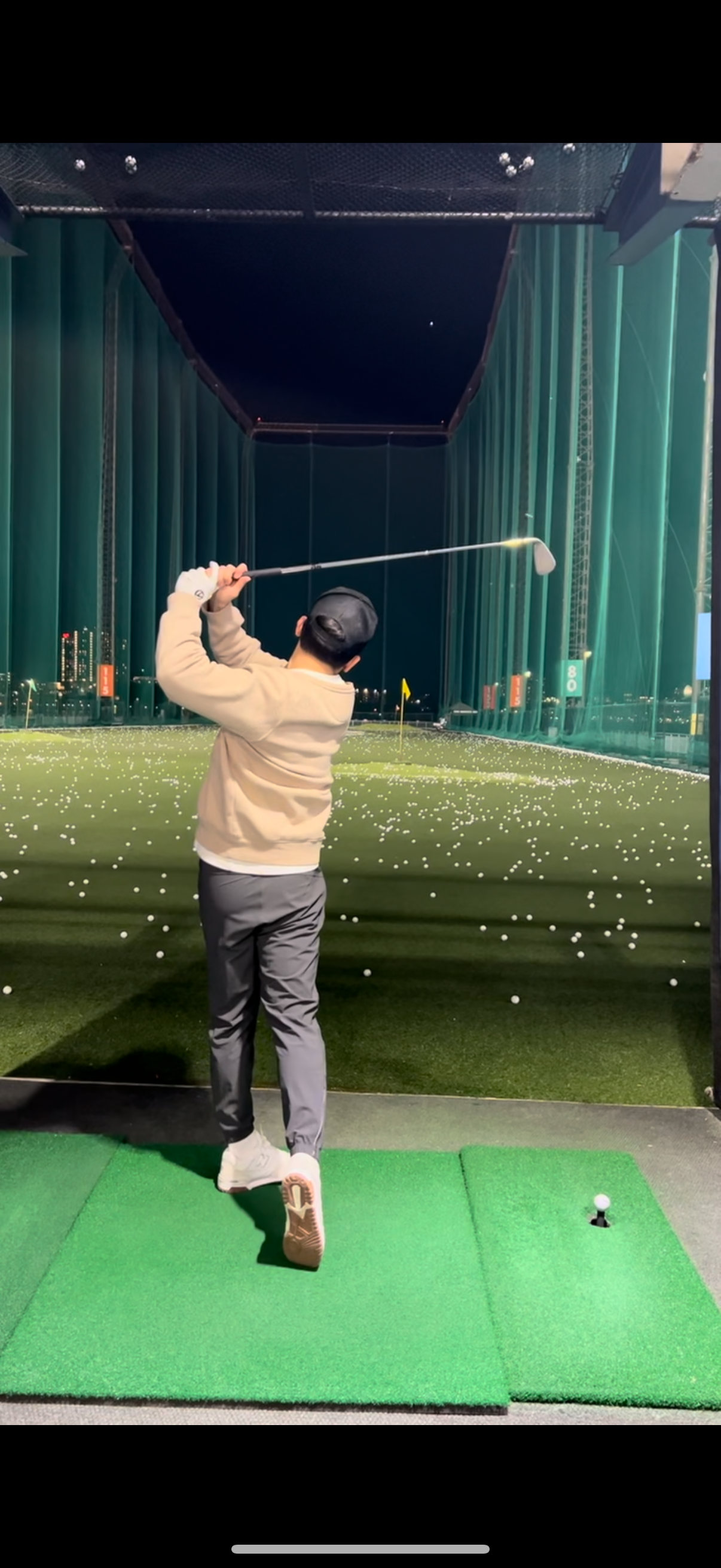 A person playing indoor golf at a driving range, swinging a golf club, with numerous golf balls scattered on the green of the range, and city lights visible in the background.