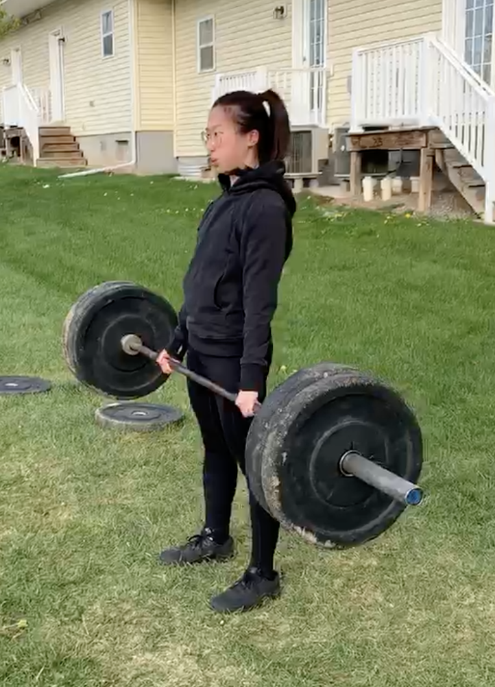 Young woman lifting a barbell with weights outdoors on a grassy yard, near a yellow house with stairs and white railing.
