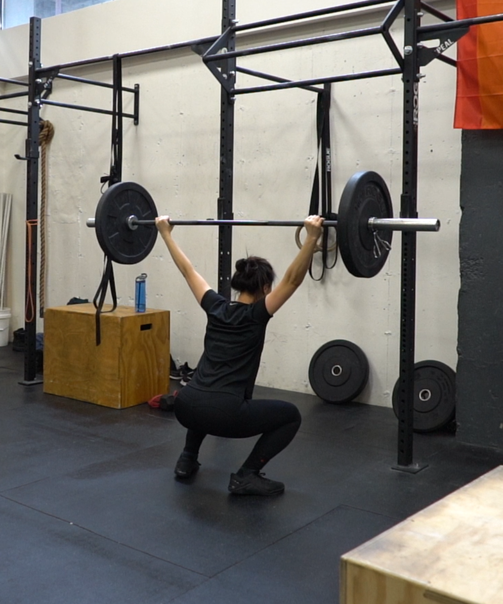A woman in black athletic clothing performing an overhead squat with a loaded barbell in a gym.