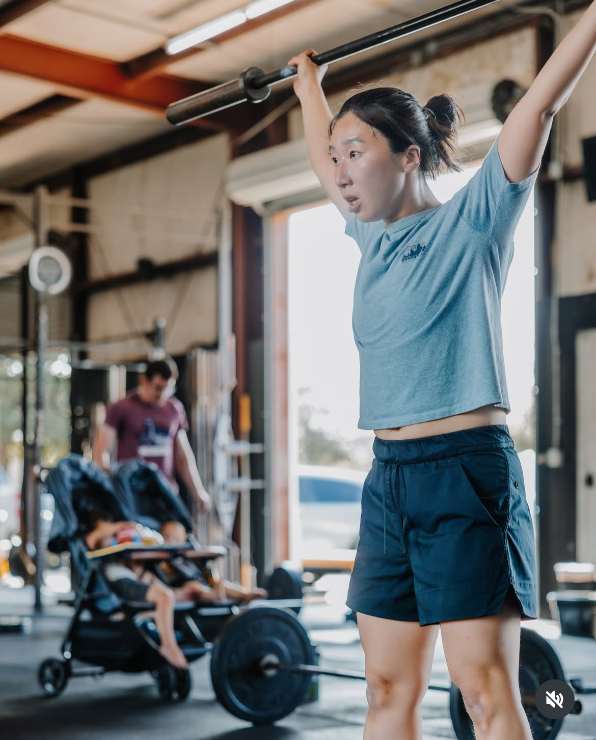 A woman lifting a barbell overhead in a gym, with a young child in a stroller and a man in the background.