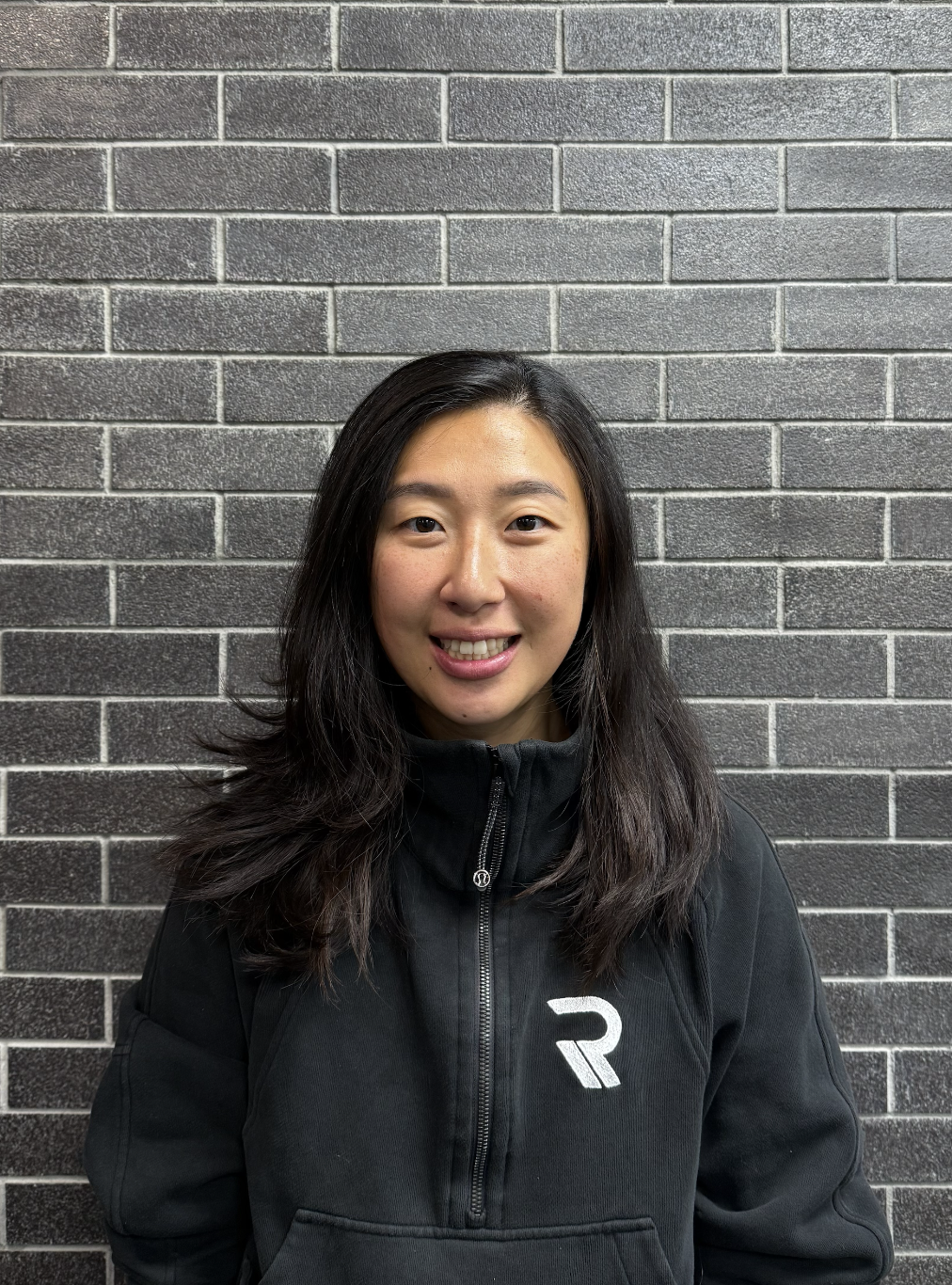 A smiling woman with long black hair wearing a black zip-up hoodie standing in front of a dark brick wall.