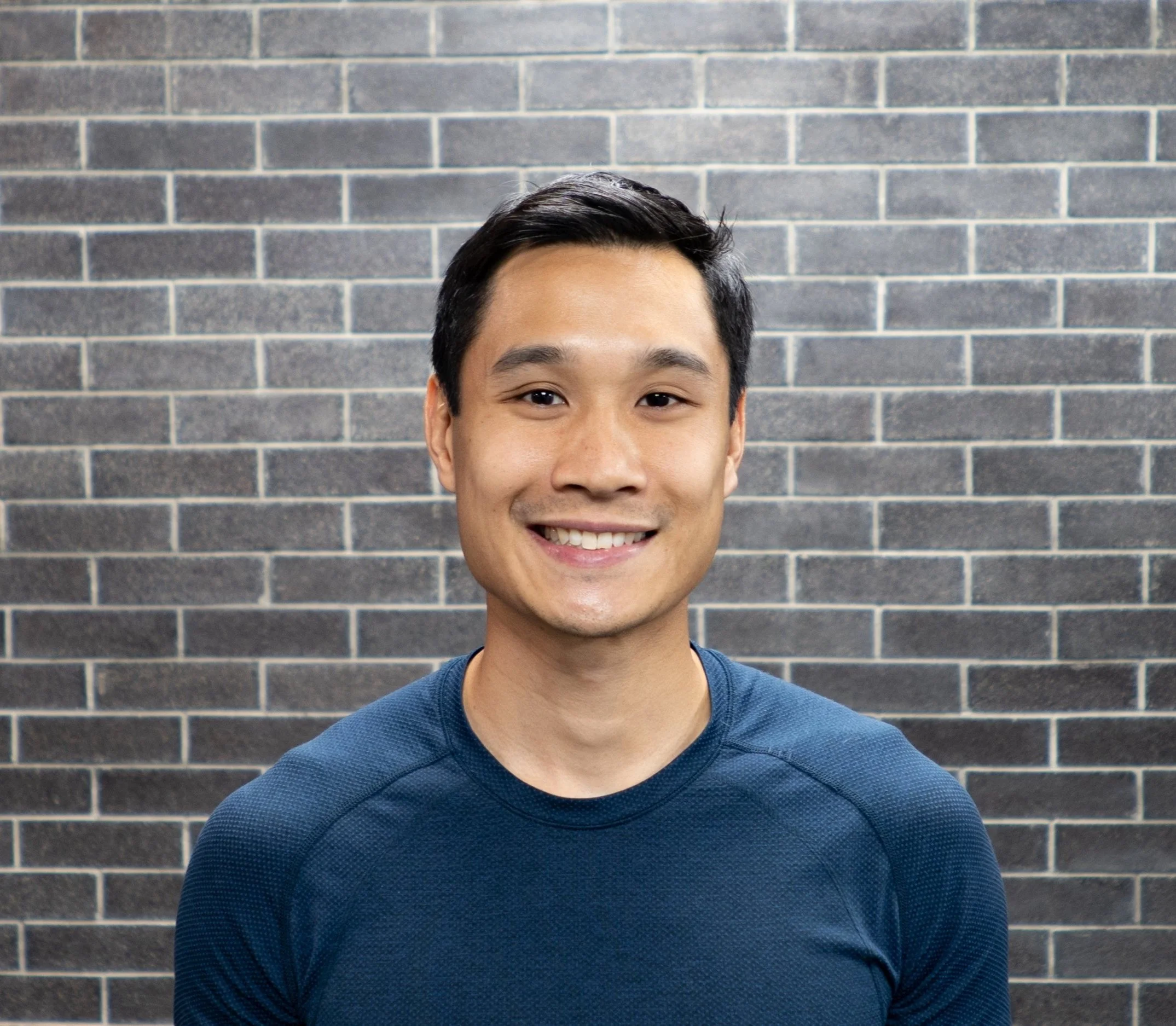 A smiling man with short black hair wearing a dark blue athletic shirt standing in front of a brick wall.