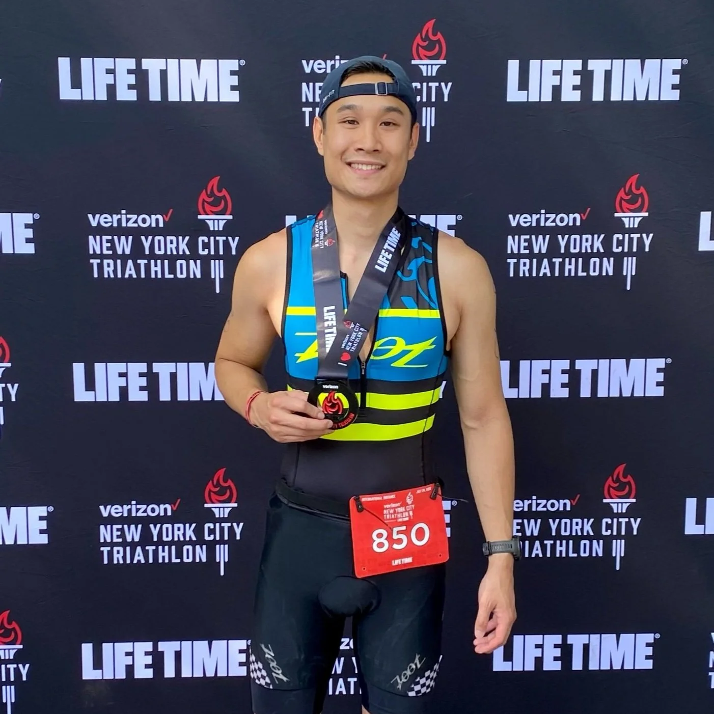 A man smiling, wearing a black cap backwards, a blue and yellow athletic skin suit, black compression shorts, and a red race bib with the number 850, standing in front of a black backdrop with logos and text from the New York City Triathlon and Verizon. He is holding a finisher medal around his neck.
